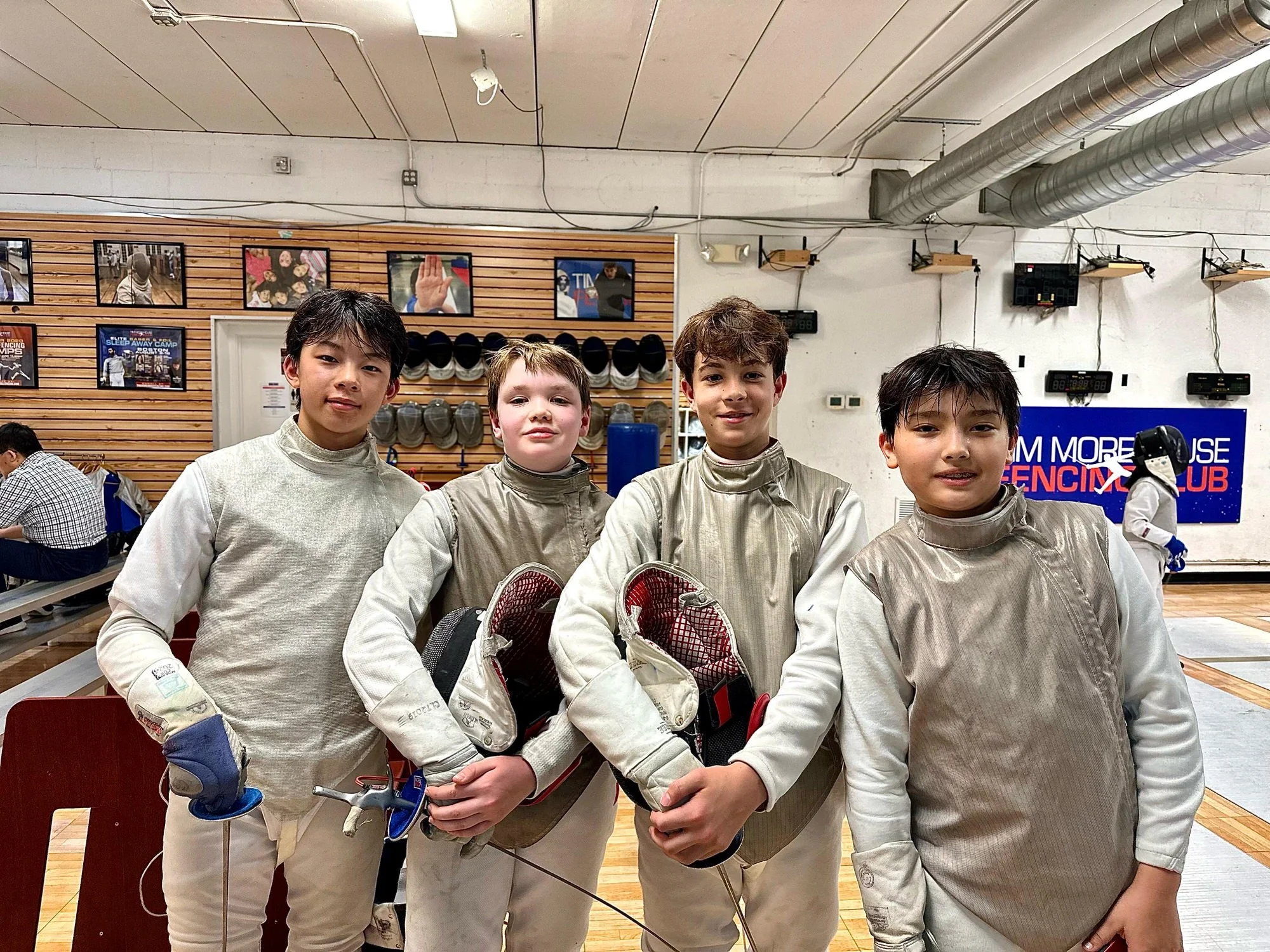 Four young boys in fencing uniforms standing together in a fencing club with one boy holding fencing masks and gear. Background includes posters, hats, and equipment on a wooden wall, with a large sign that says 'FENCING CLUB' in the background.