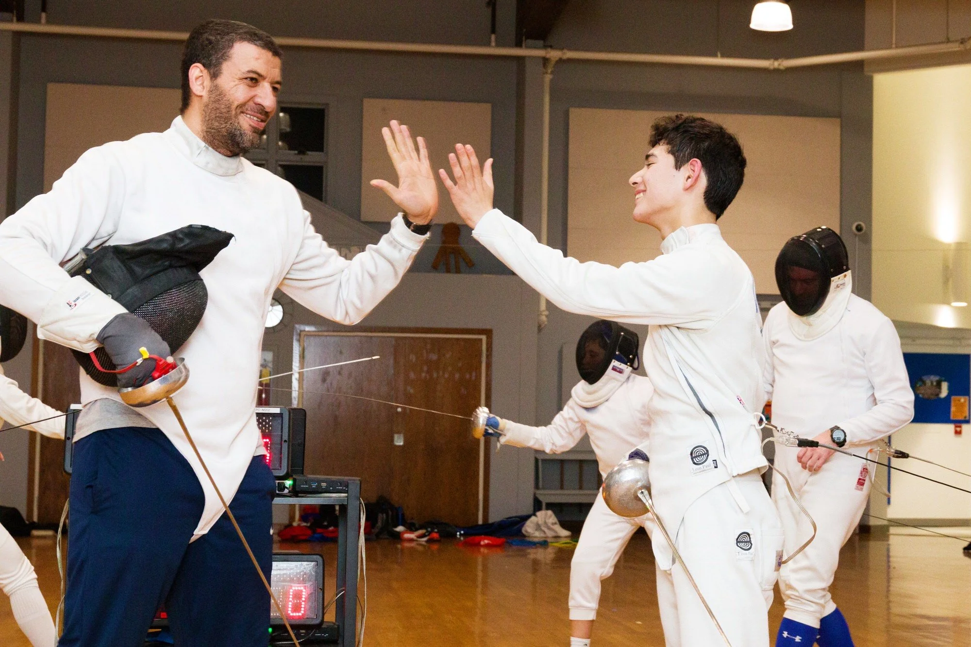 A fencing coach and a smiling male fencer exchange a high five during a fencing practice or class in an indoor gym, with other fencers and fencing equipment visible in the background.