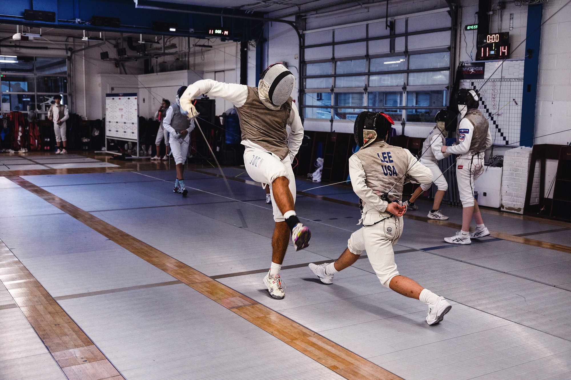 Two men engaged in a fencing duel with swords in an indoor fencing gym, surrounded by other people practicing and observing.