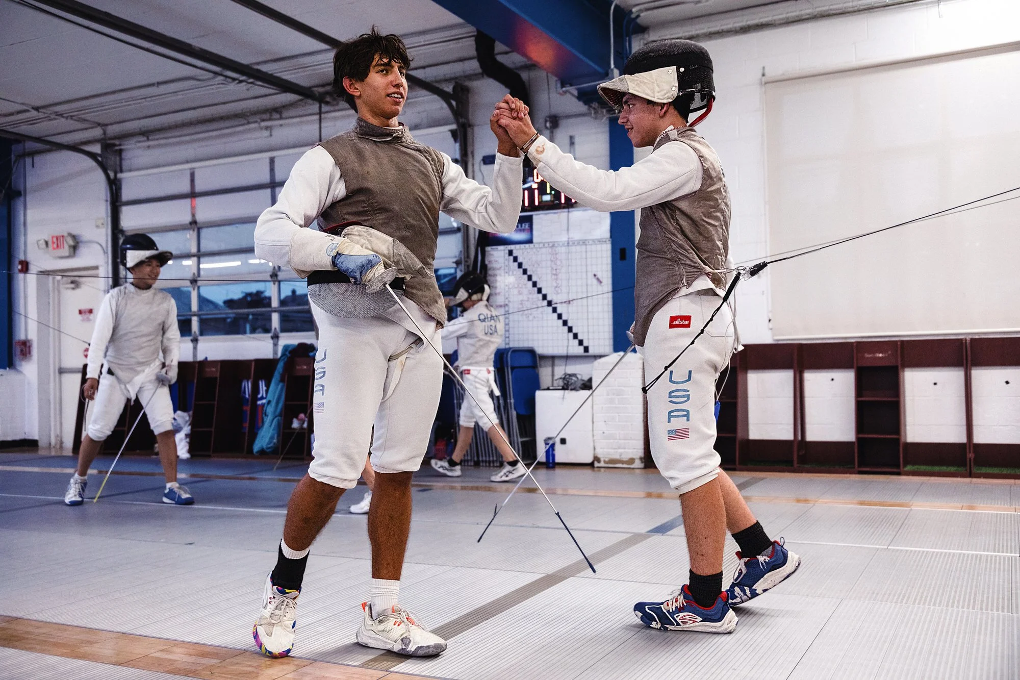 Two male fencers in fencing gear celebrate with a high five in a fencing gym, with two other fencers in the background preparing.