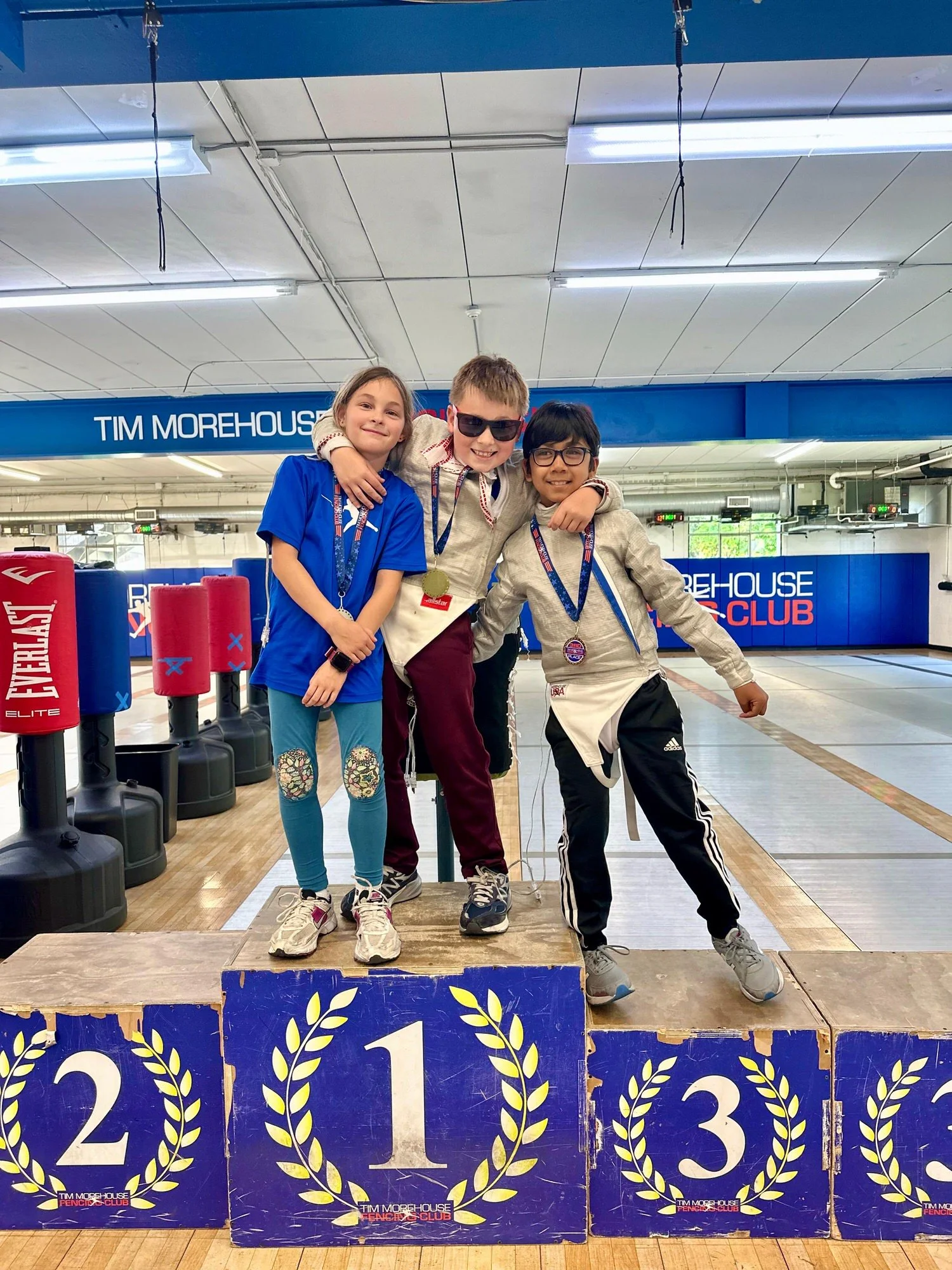 Three children standing on a winners' podium in a fencing club, holding medals and smiling. The first-place winner in the center has their arms around the second and third-place winners. The backdrop shows fencing equipment and a banner with text.