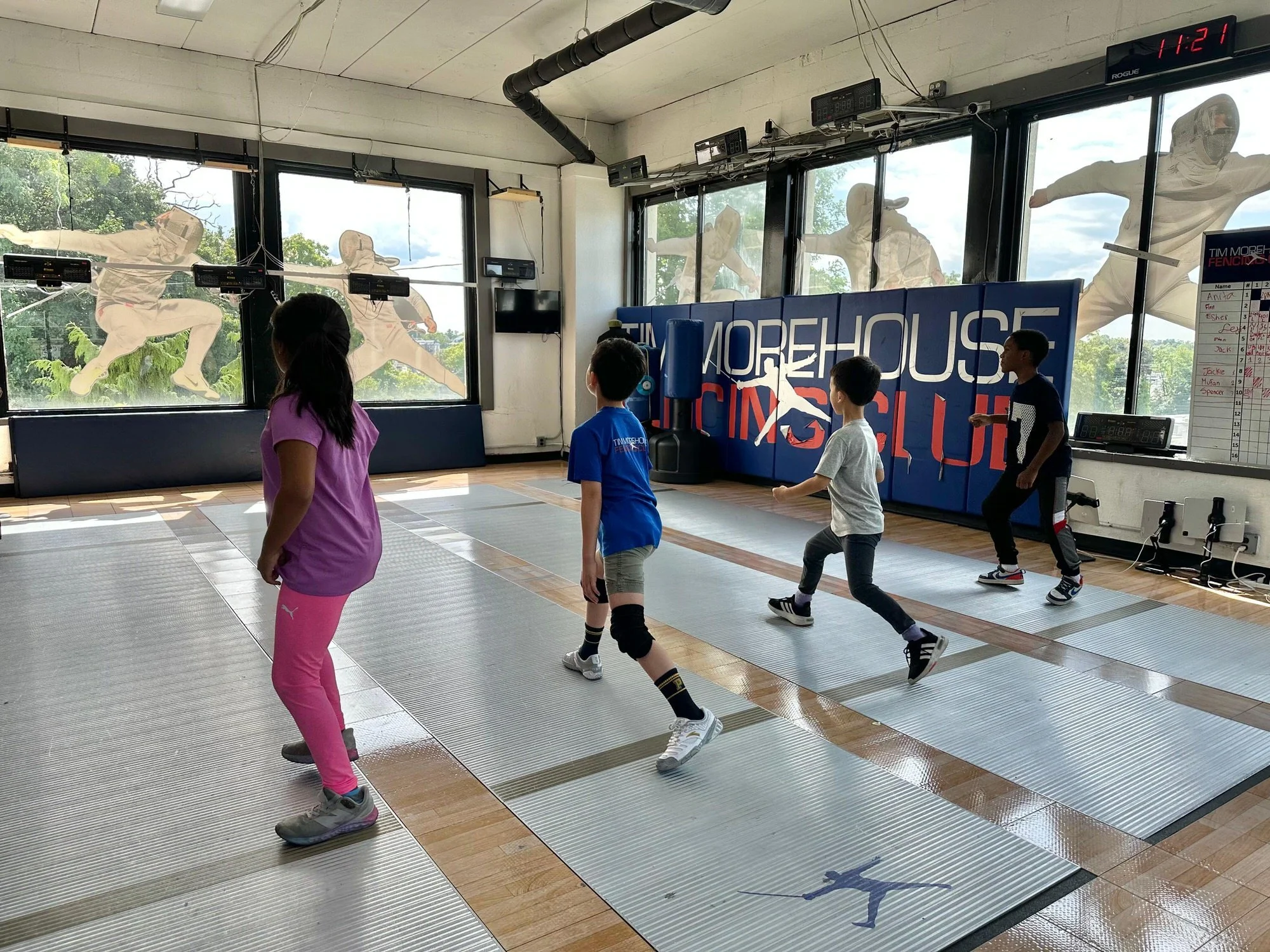 Children in a fencing class on padded mats inside a gym, with large windows displaying athletic figures in fencing poses on the exterior, and a digital clock on the wall showing 11:21.