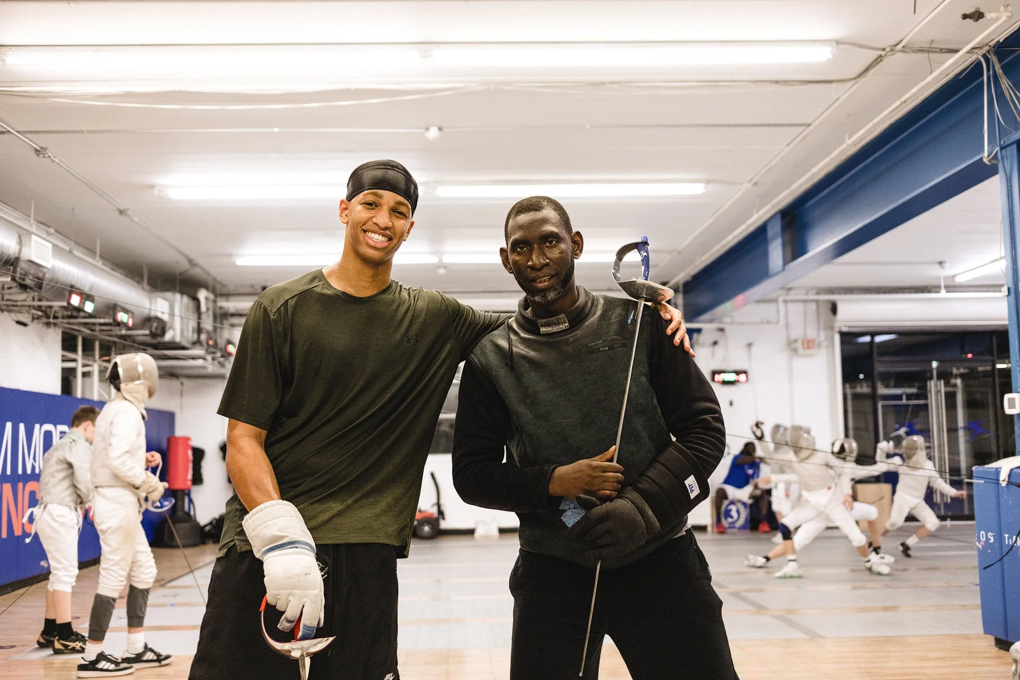 Two men standing in an indoor fencing training facility, smiling, with fencing equipment and other fencers practicing in the background.