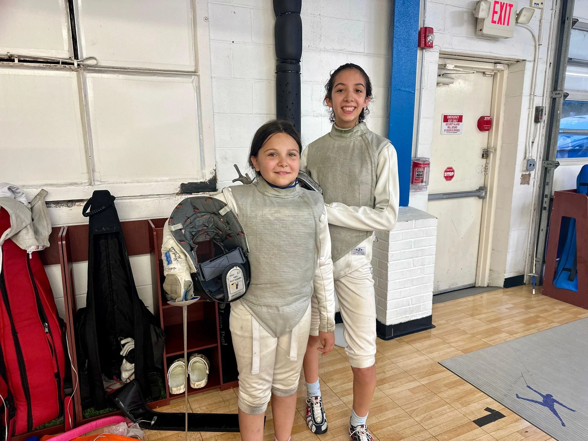 Two young female fencers in fencing gear smiling indoors, standing near a white brick wall and fire emergency equipment. One girl holds a fencing mask, and they are dressed in protective fencing attire.