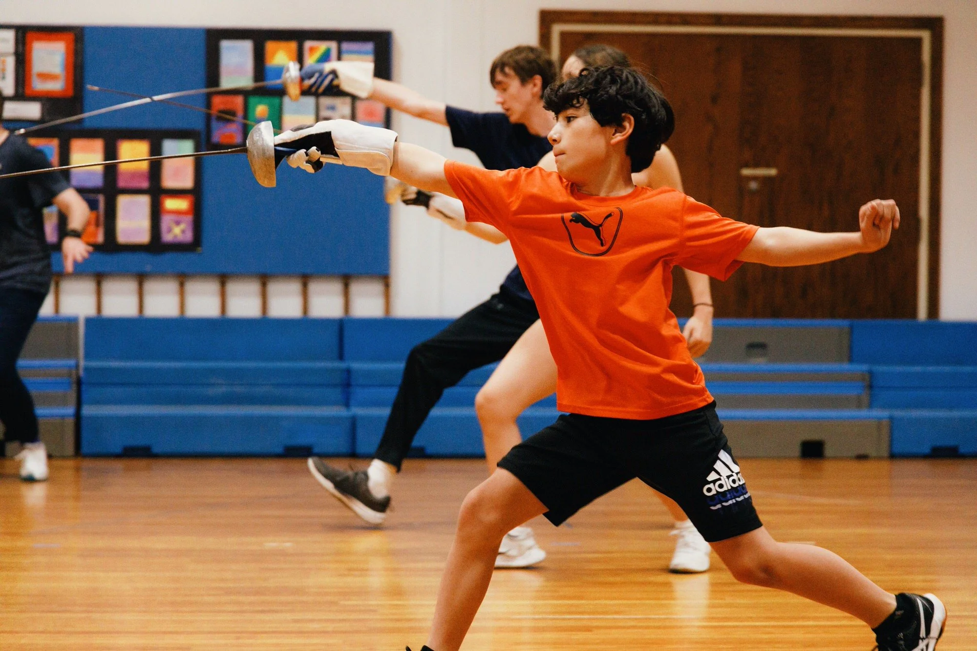 Children practicing fencing in a gymnasium, with wooden floors and colorful artwork on a bulletin board in the background.