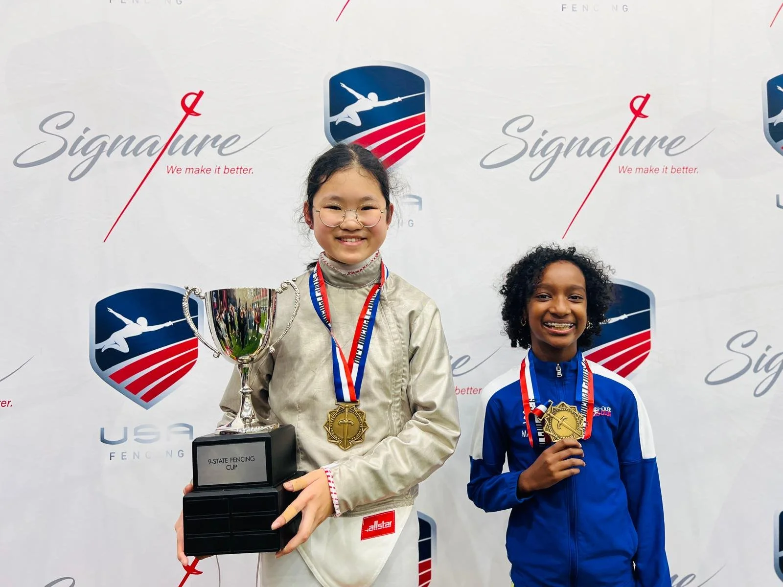 Two young girls standing in front of a backdrop with logos, smiling, holding medals and a trophy, celebrating a fencing competition.