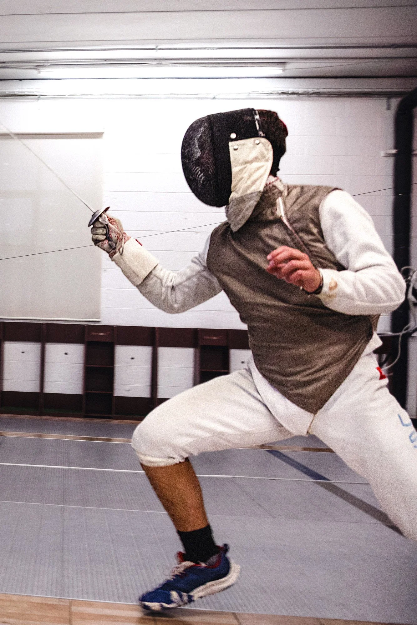 A person practicing foil fencing in a gym, wearing protective fencing gear, including a mask and jacket, and holding a foil sword in a fencing stance.
