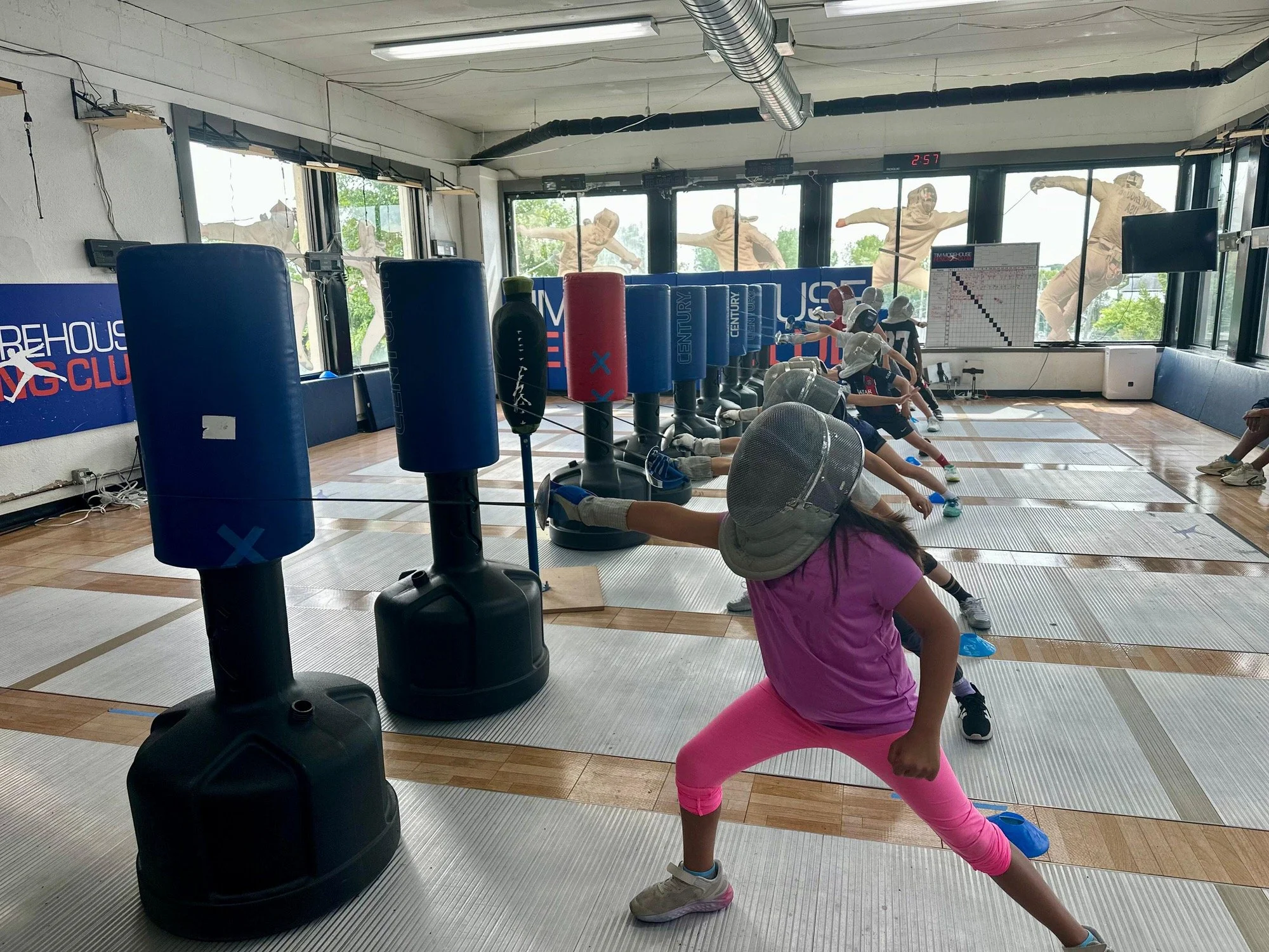 Children participating in a fencing workout at a gym with punching bags, mats, and a large mirror, outside natural light coming through windows.