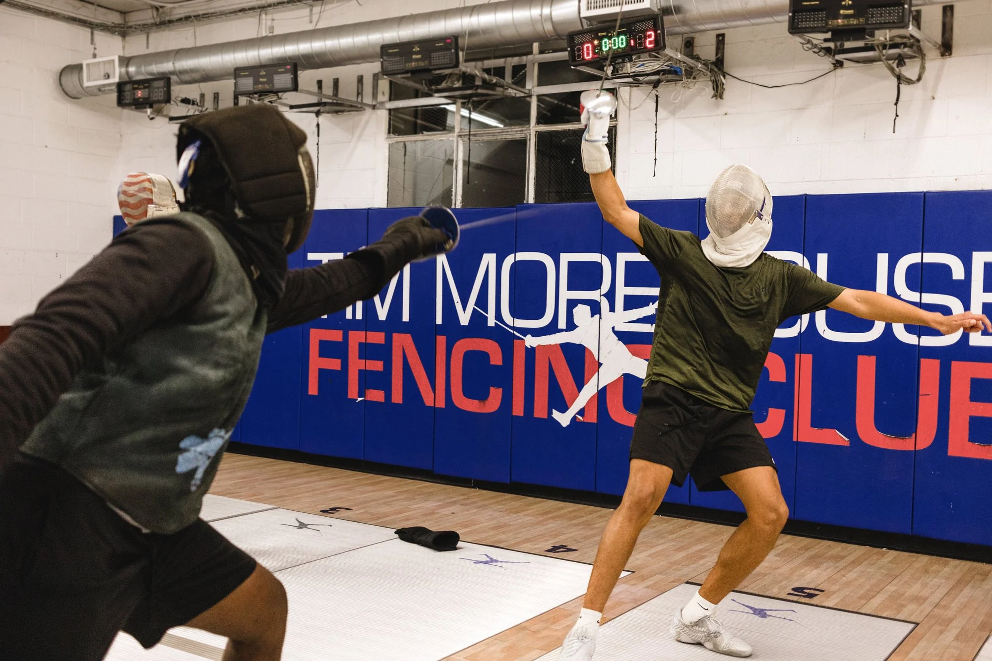 Two fencers practicing with masks and protective gear in a gym, one leaping with a weapon aiming at the other, with a fencing club sign in the background.