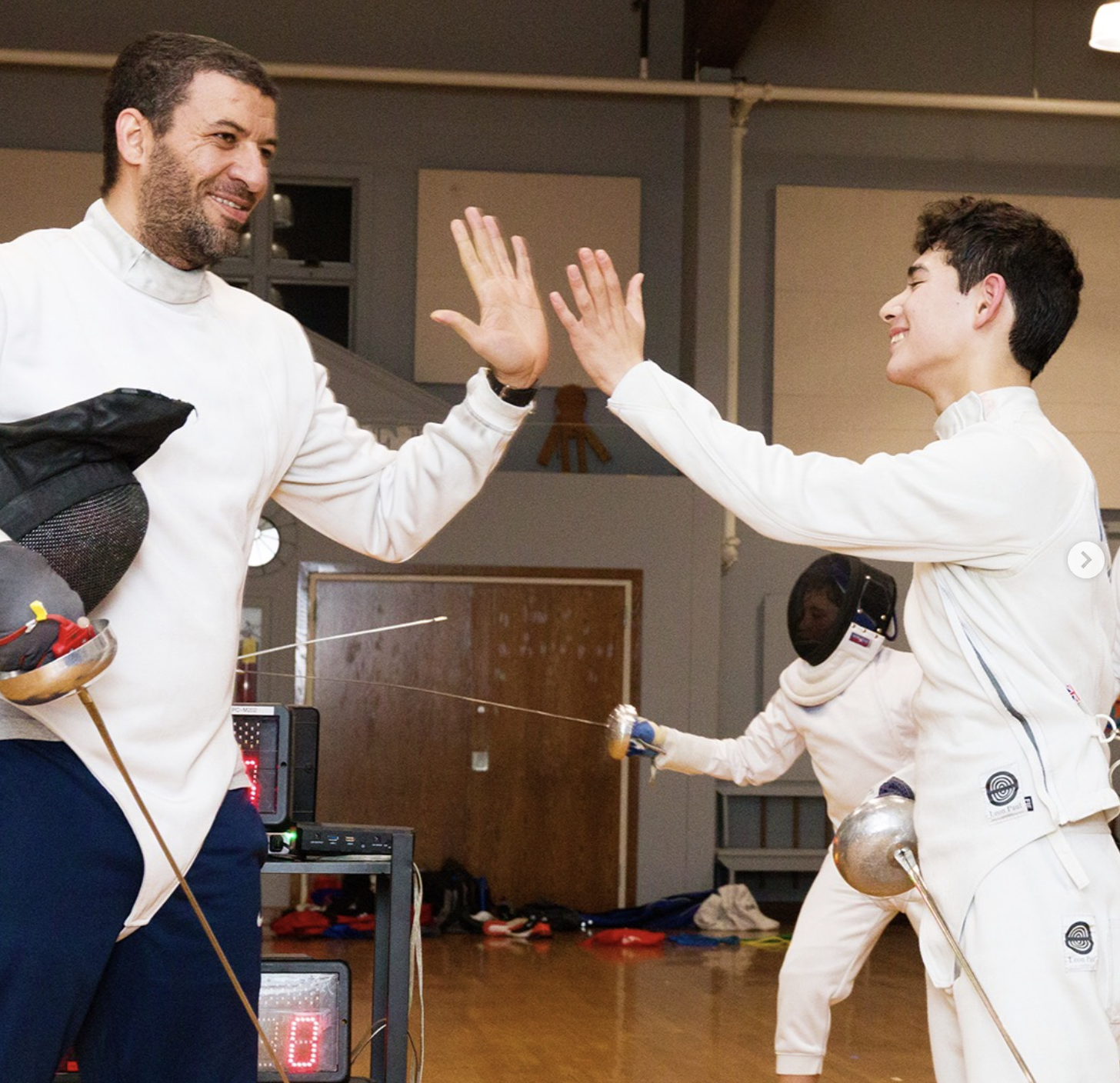 Young boy practicing fencing with an adult coach inside a gym, with large windows showing a cloudy sky outside.