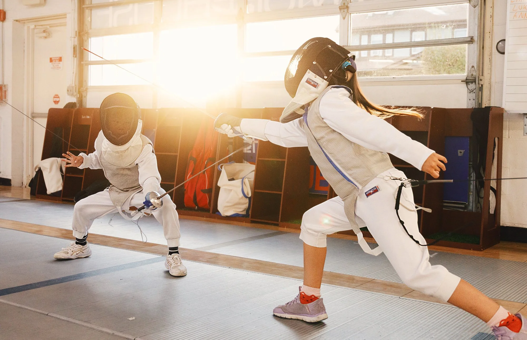 Two children practicing fencing indoors during sunset.