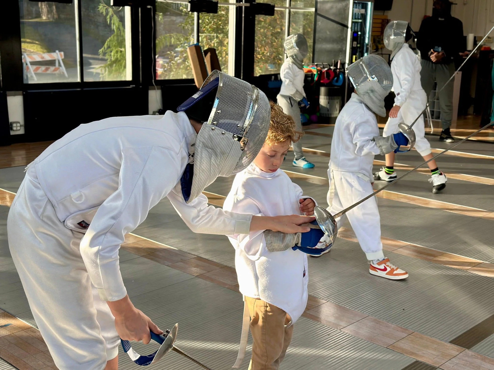 Children dressed in fencing gear practicing fencing in a gym with large windows.