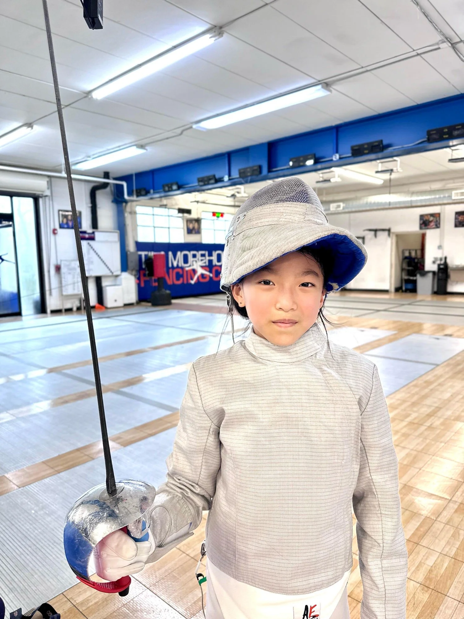 Young girl in fencing gear holding a fencing sword in a fencing gym with mats on the floor and fencing equipment in the background.