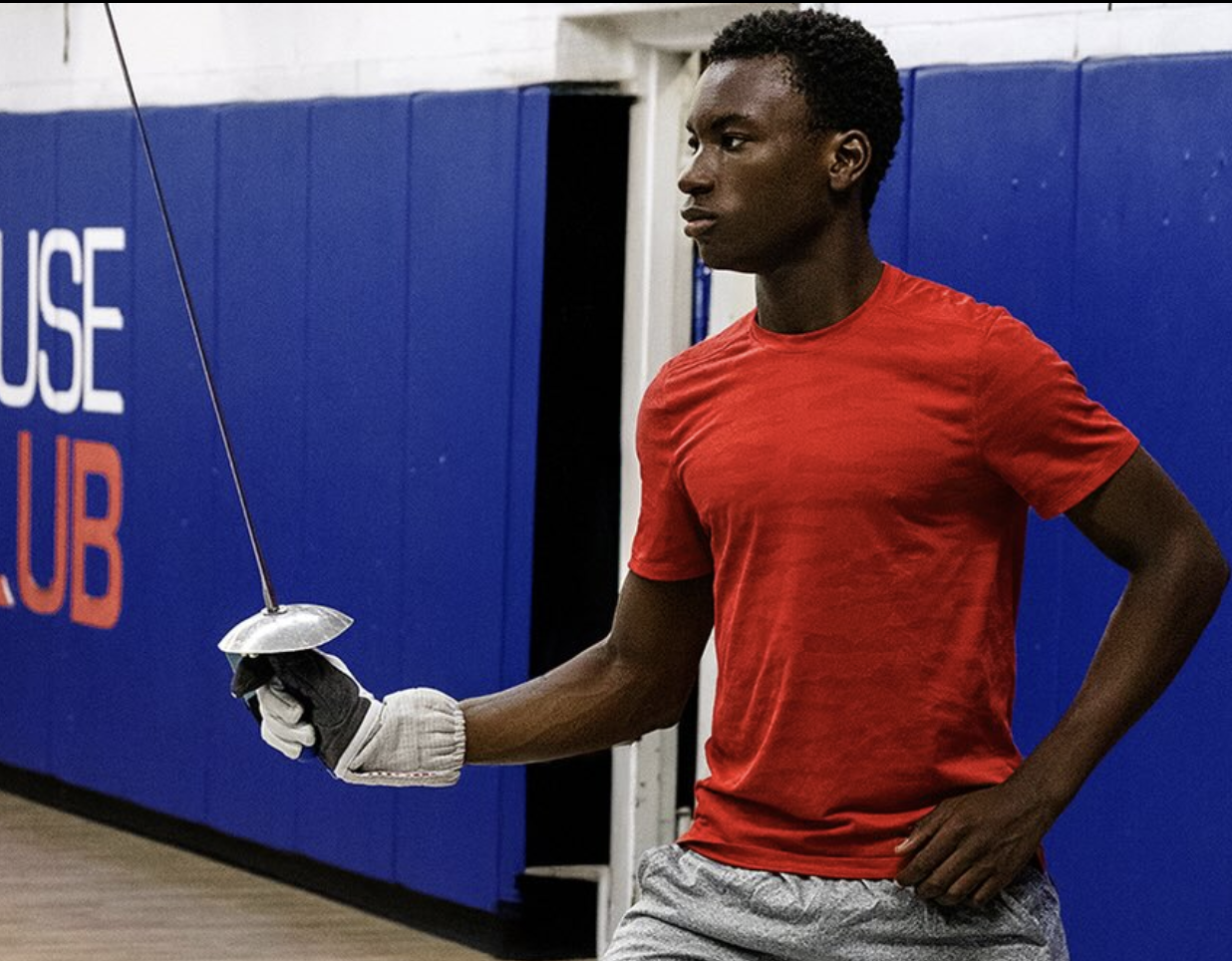 Children dressed in fencing gear posing for a photo in a fencing gym, with other people fencing in the background.