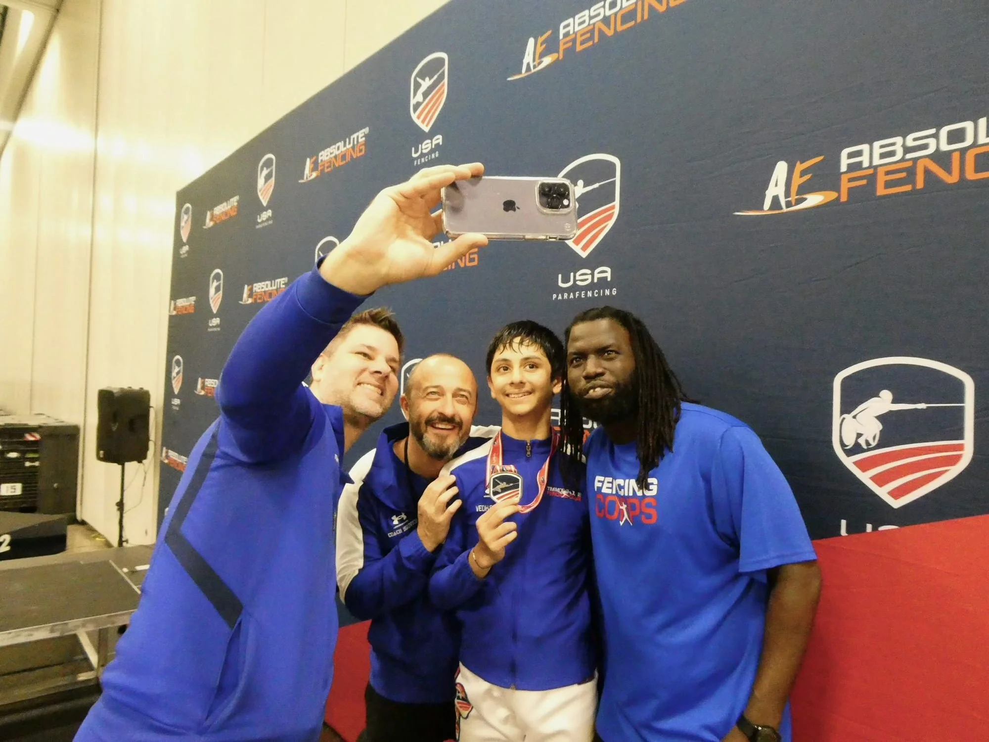 Four men taking a selfie at a USA parafencing event, with medals around their necks, standing in front of a blue backdrop with logos of USA fencing and Absolute Fencing.