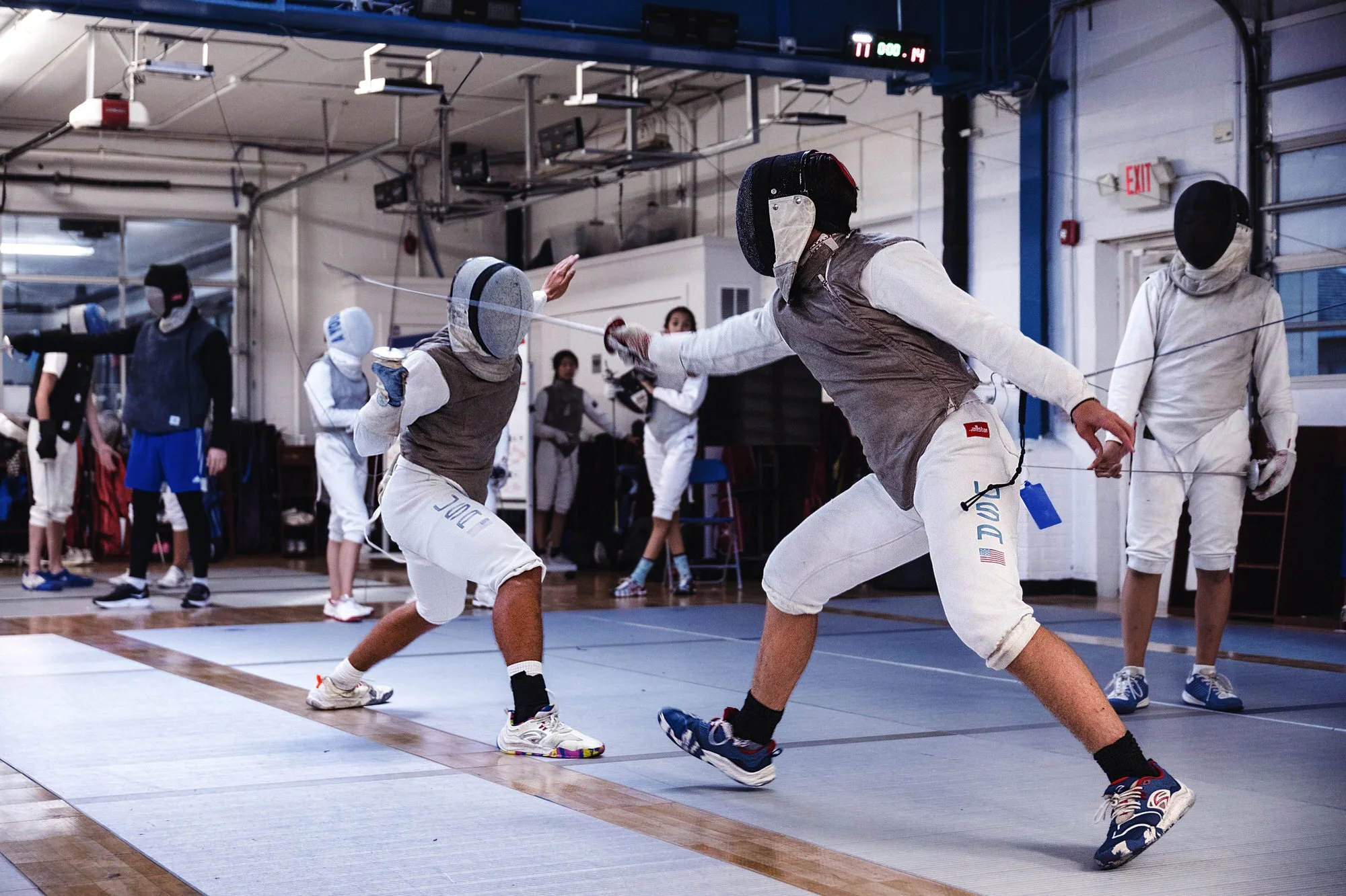 Two fencers in white and brown protective gear engaged in a fencing match, facing each other on a fencing strip with other people observing in the background.