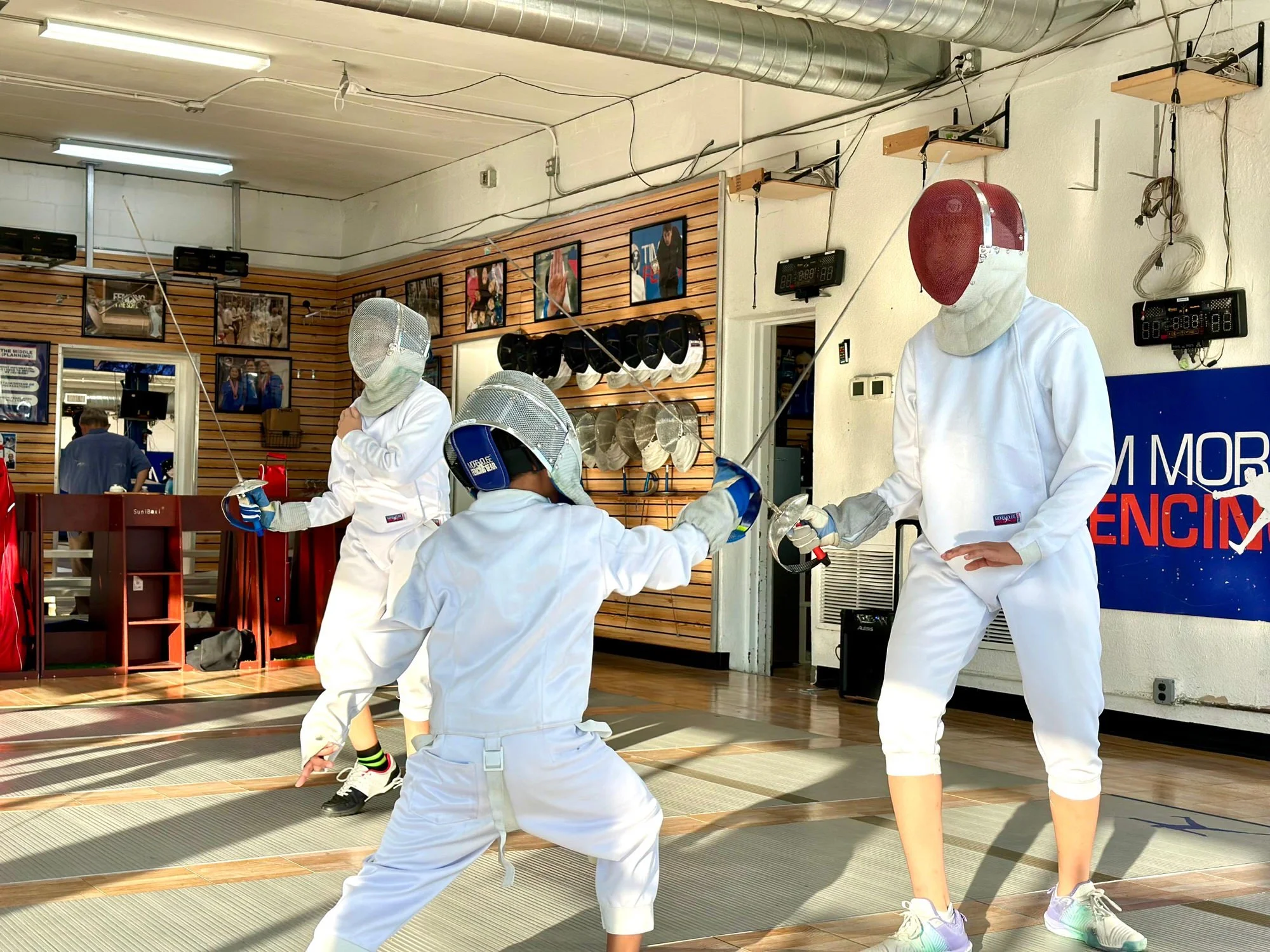 Three children practicing fencing indoors, wearing white protective gear and masks, with one child in a stance ready to duel, and an instructor in the background.
