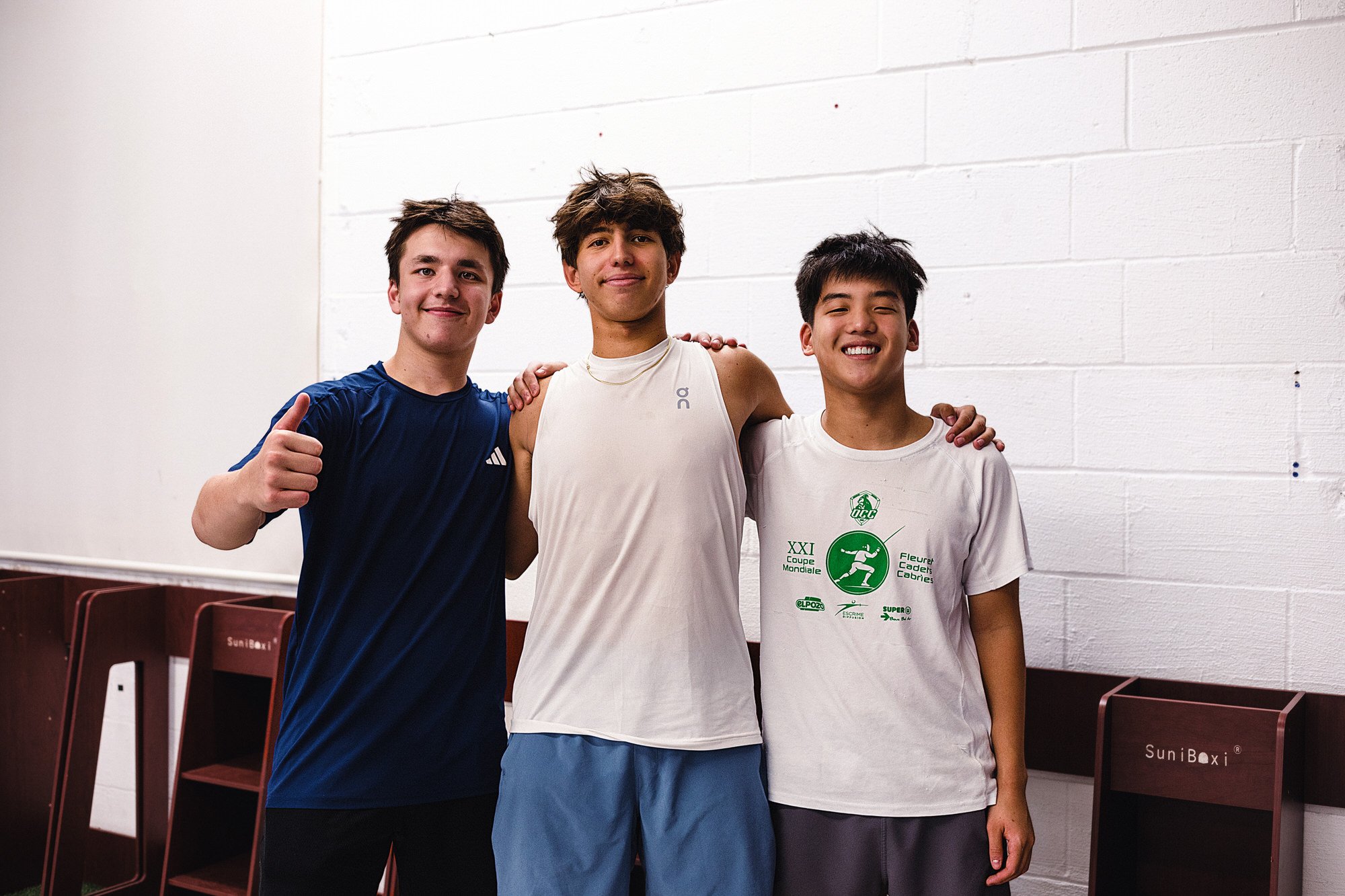 Three young male athletes standing together in a gym, smiling and with arms around each other, wearing sportswear.