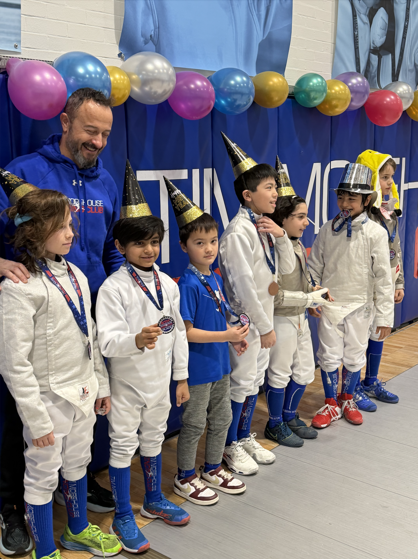 Birthday party medal ceremony at Tim Morehouse Fencing Club - kids in fencing gear and party hats with a balloon arch backdrop