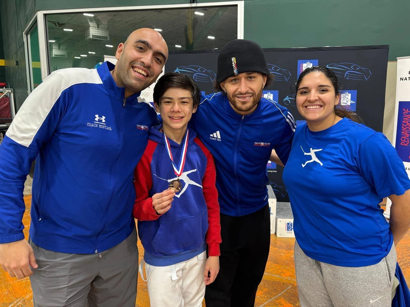 Four smiling people, two adults and two youths, standing together indoors. The boy in the center holds a gold medal, and a woman is wearing a blue shirt with a white logo. The background includes banners, one with a blue and white design. The group l