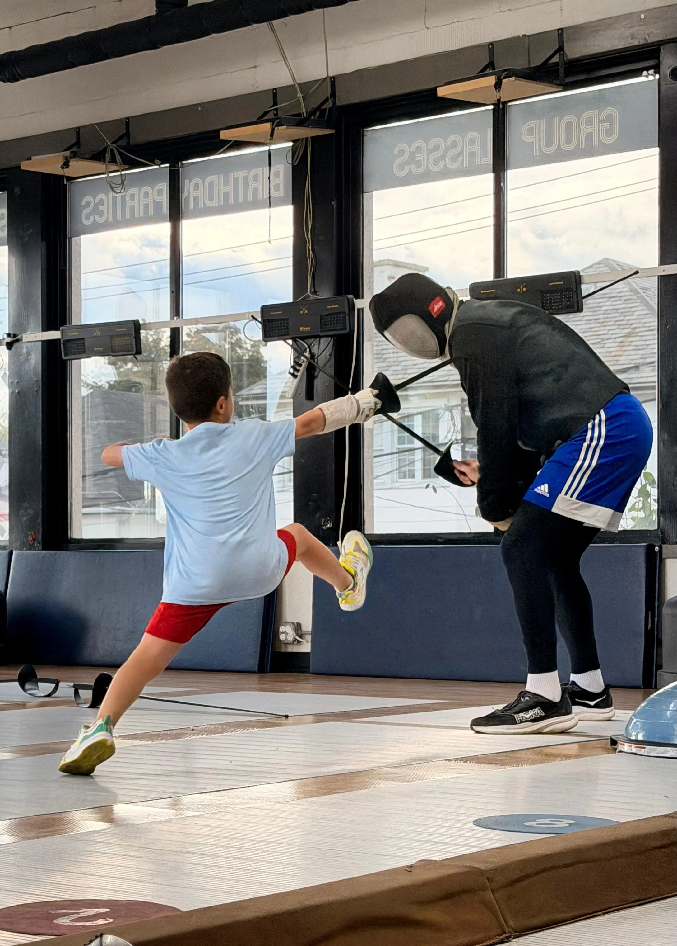 Young boy practicing fencing with an adult coach inside a gym, with large windows showing a cloudy sky outside.