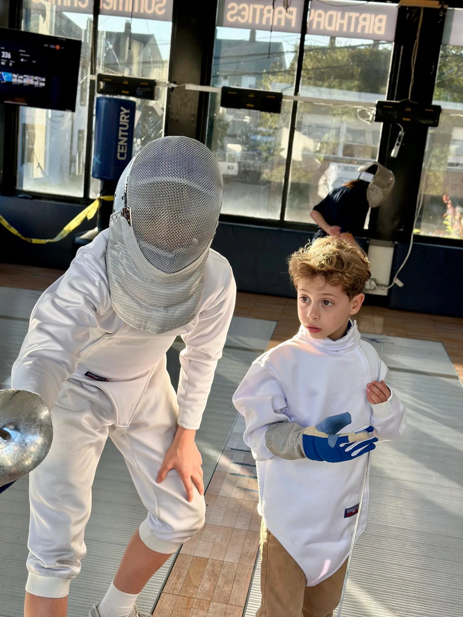 Two children in fencing gear, one adult fencing, inside a room with large windows and a sign reading 'BIRTHDAY PARTIES'