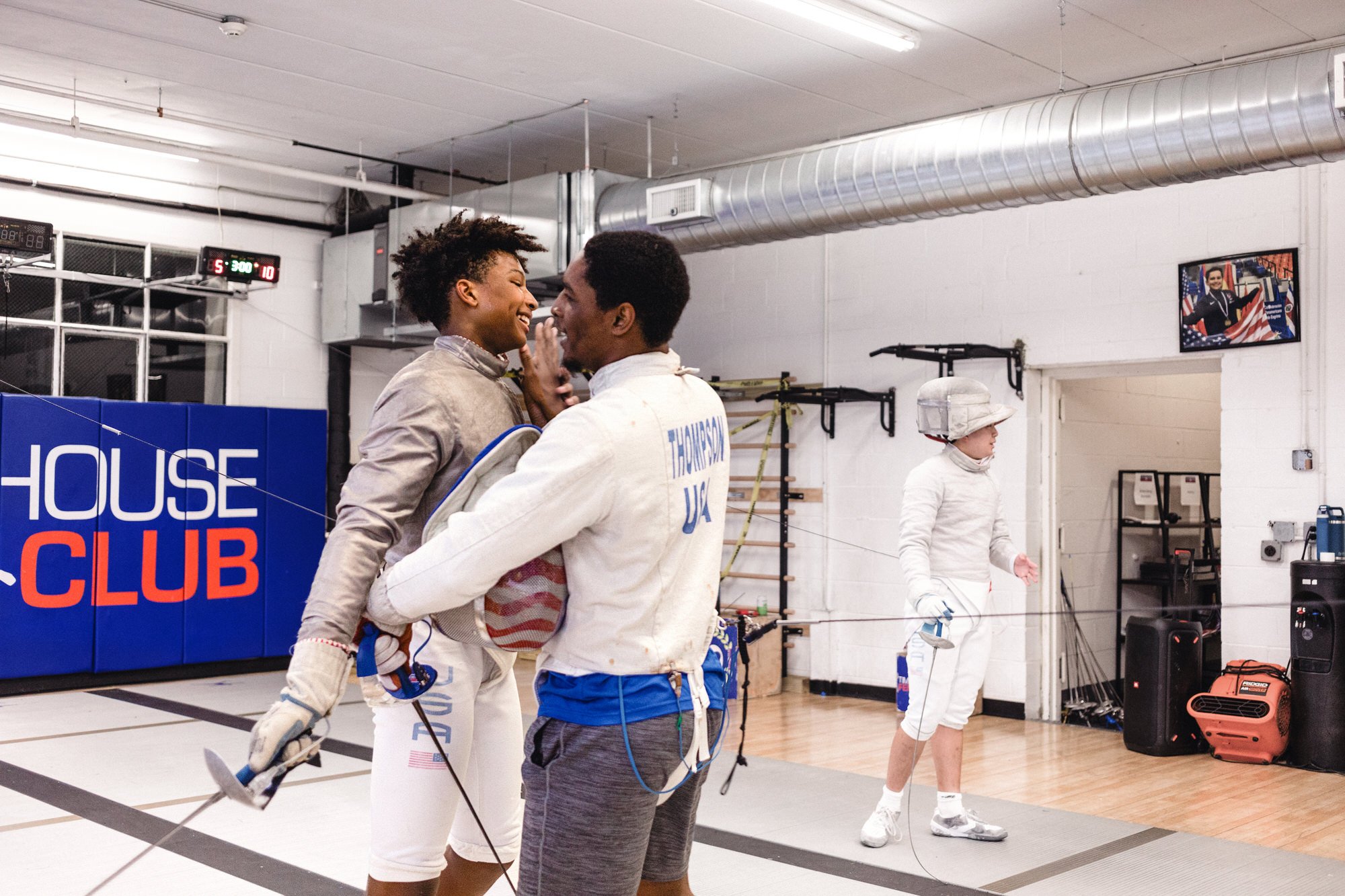 Two fencers, a woman and a man, share a joyful moment in a fencing gym, with a woman practicing fencing in the background. The gym has a sign reading 'HOUSE CLUB' and various fencing equipment on the walls.