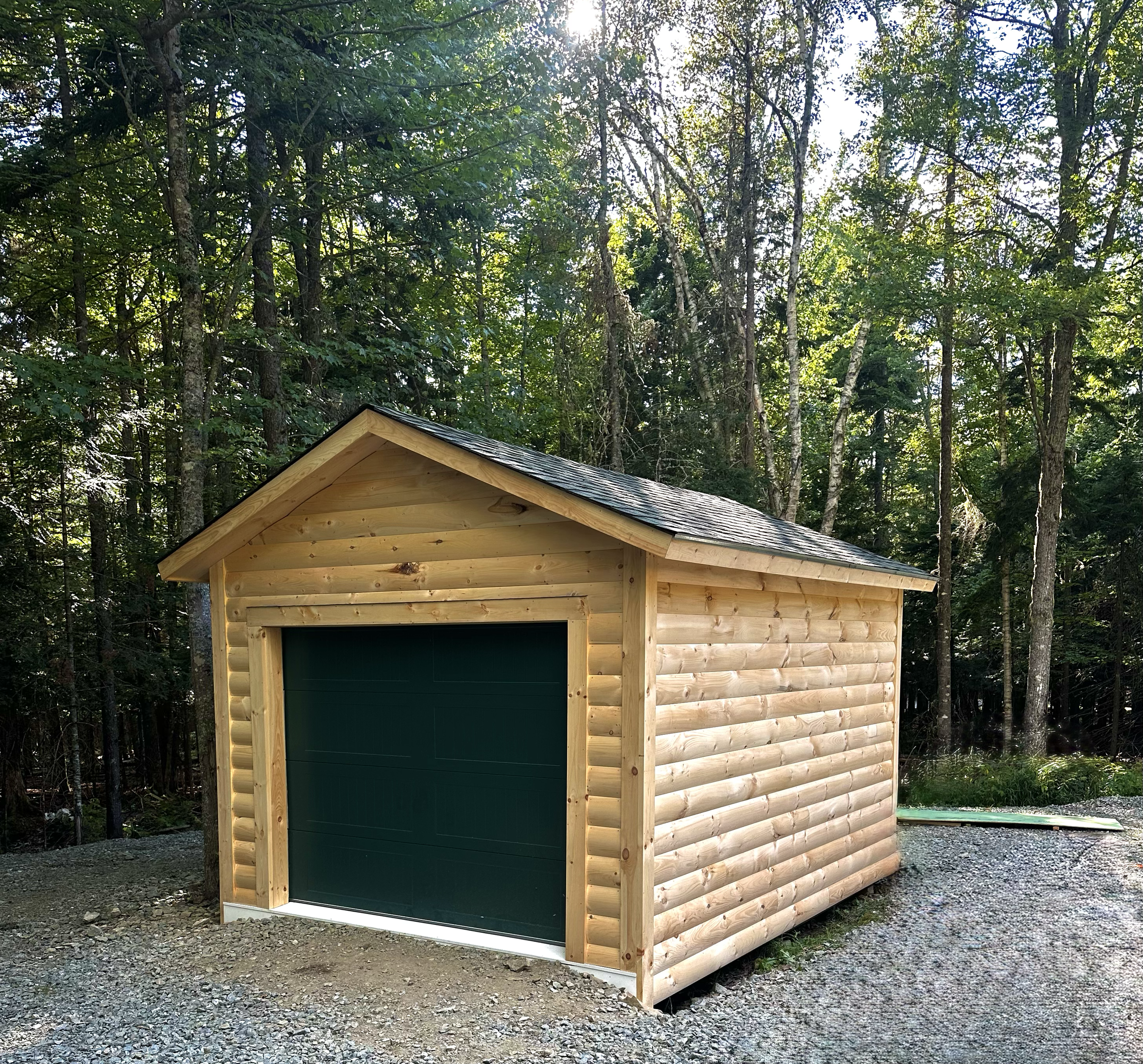 A small wooden shed with a green garage door, situated outdoors on gravel, surrounded by tall trees and greenery.