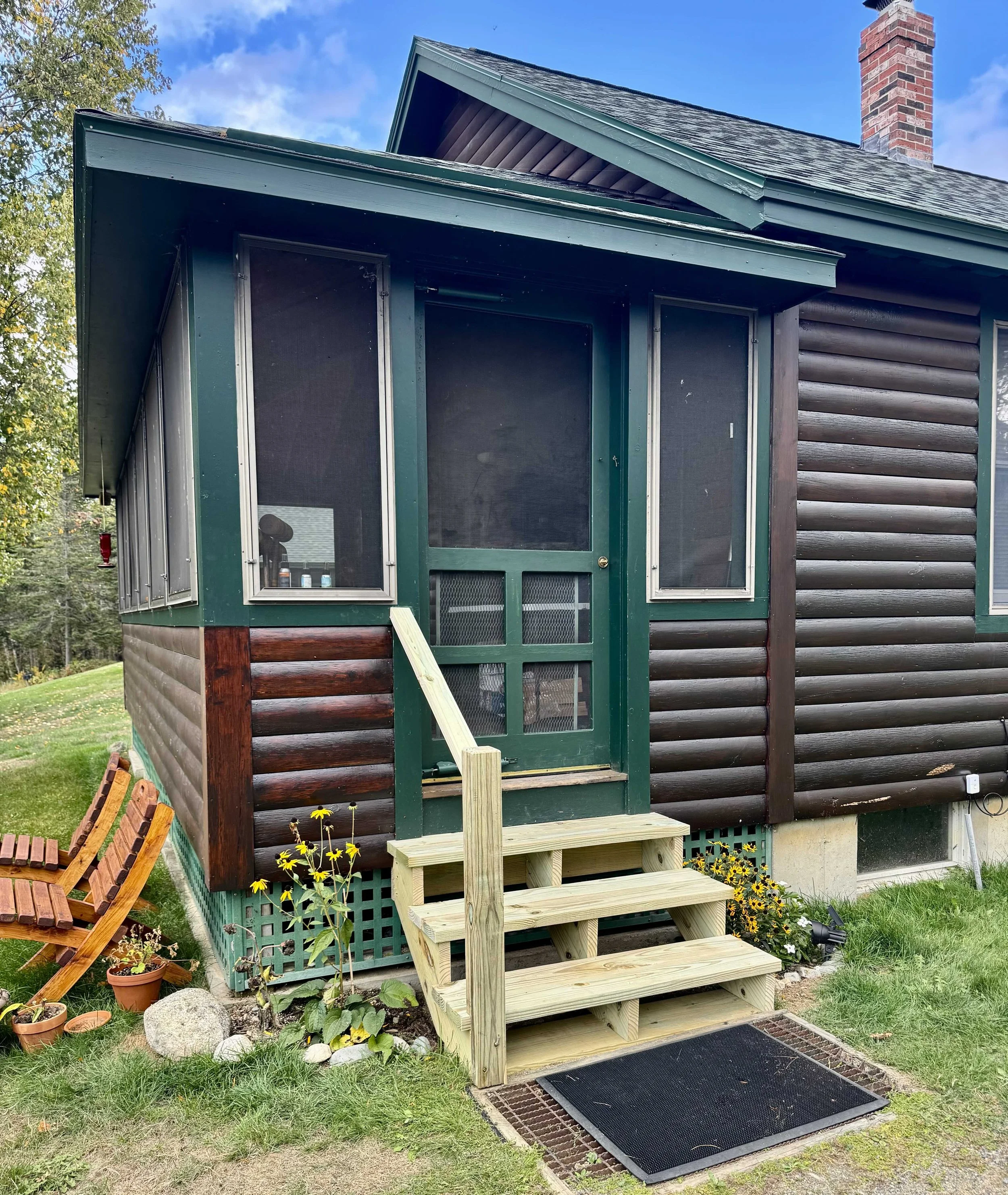 Front porch of a house with a small staircase, screened door, and windows, surrounded by green grass, flowers, and trees, under a blue sky with clouds.