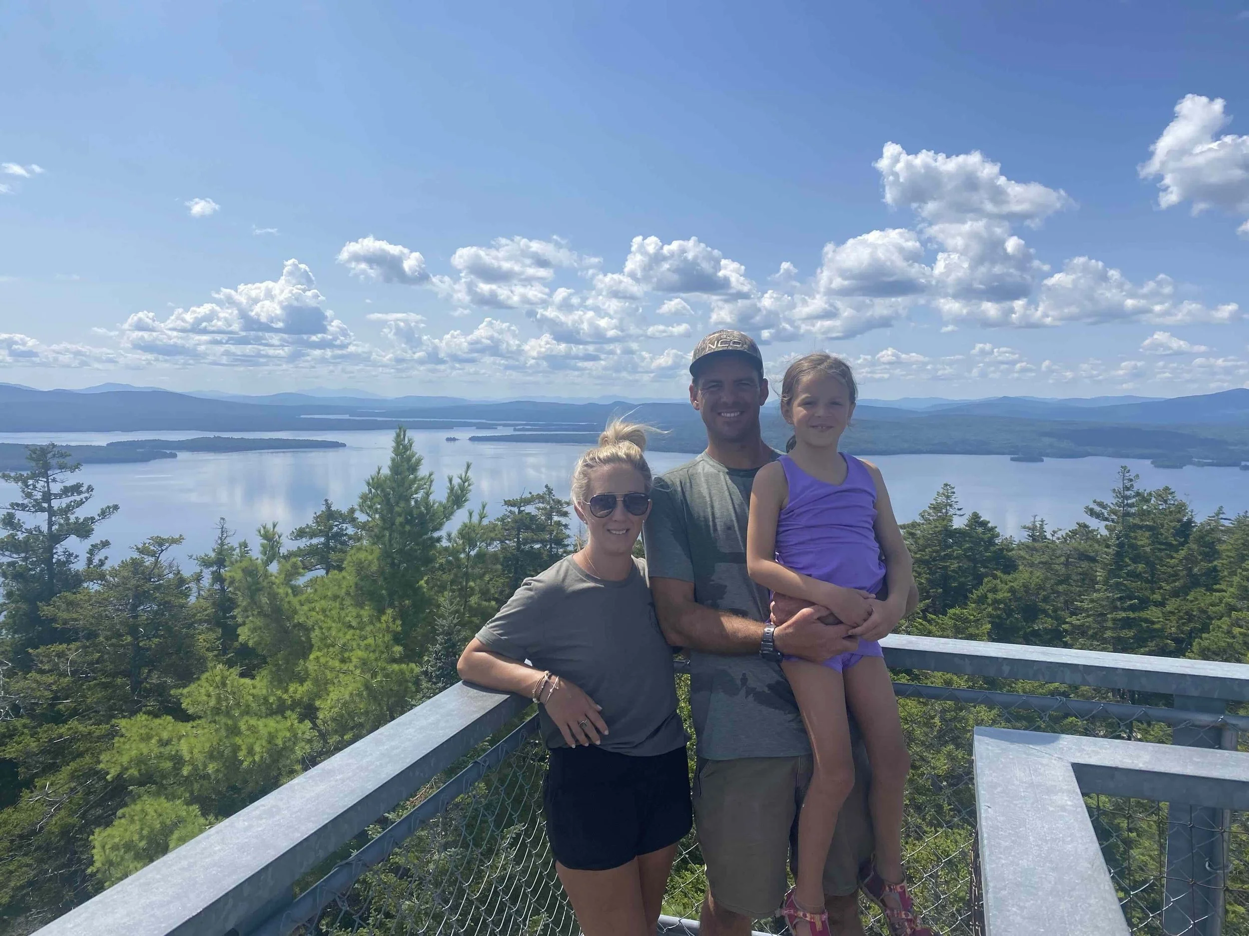 A family of three standing on an observation deck overlooking a lake surrounded by trees and mountains on a sunny day with scattered clouds.