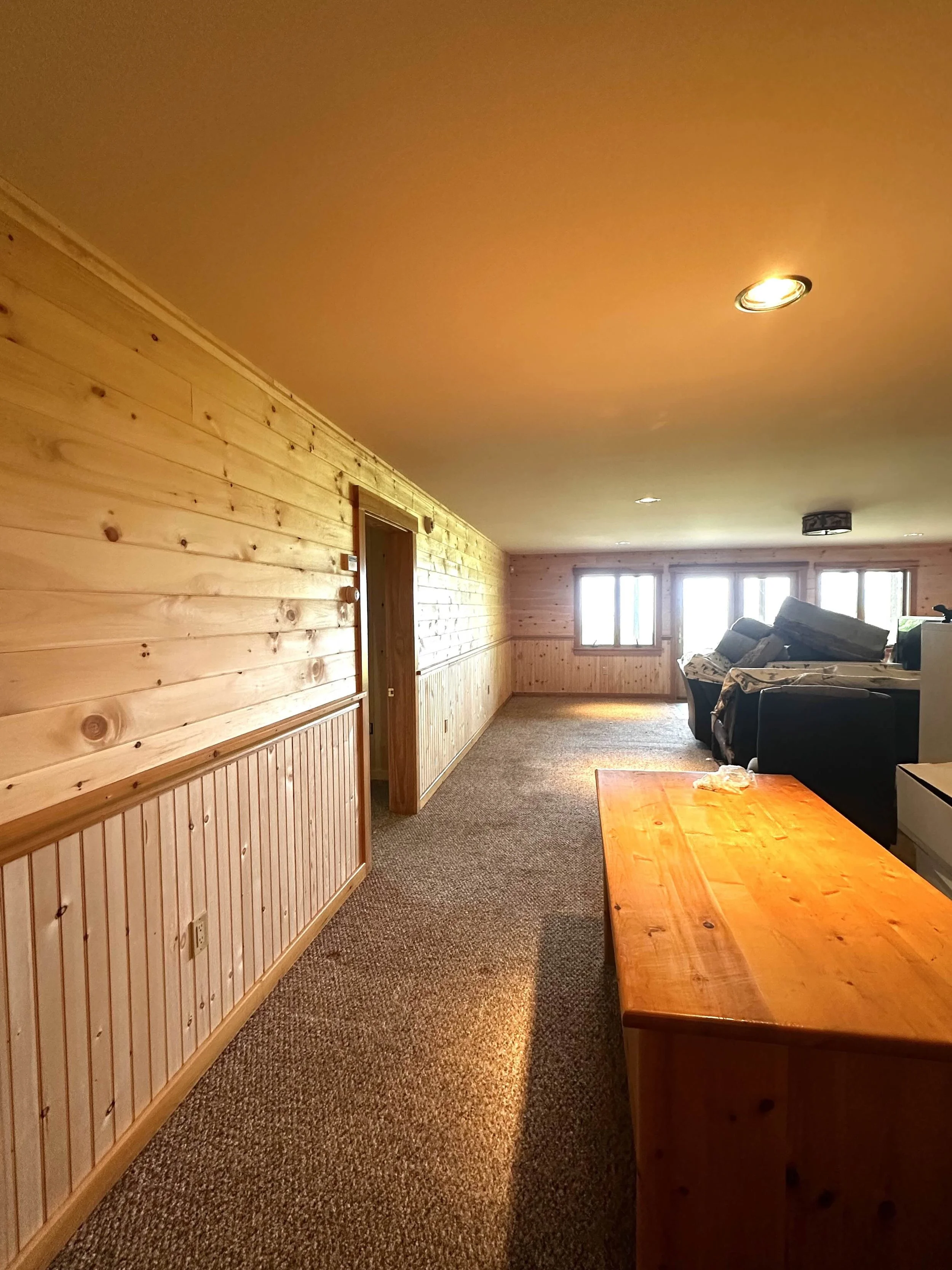 A spacious living room with wood-paneled walls, carpeted floor, and a row of windows at the far end letting in natural light. There are stacked furniture pieces near the windows and a simple wooden table in the foreground.