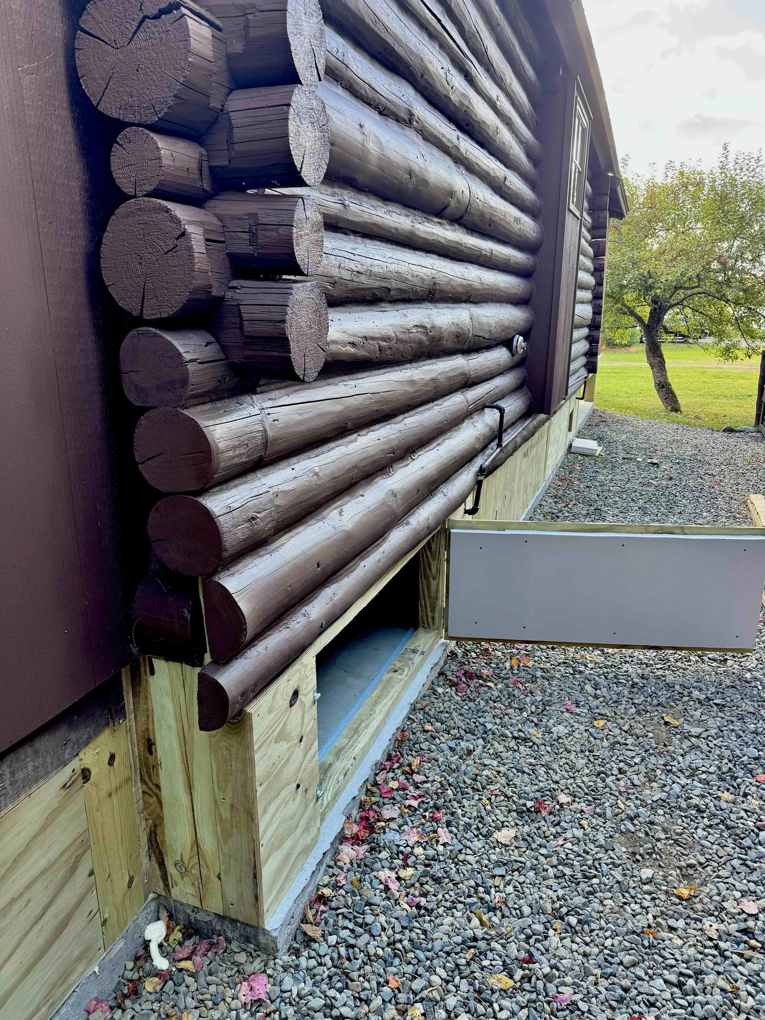 Close-up of a log cabin exterior with brown wooden logs and a small white vent, situated on gravel ground with a tree and grass in the background.