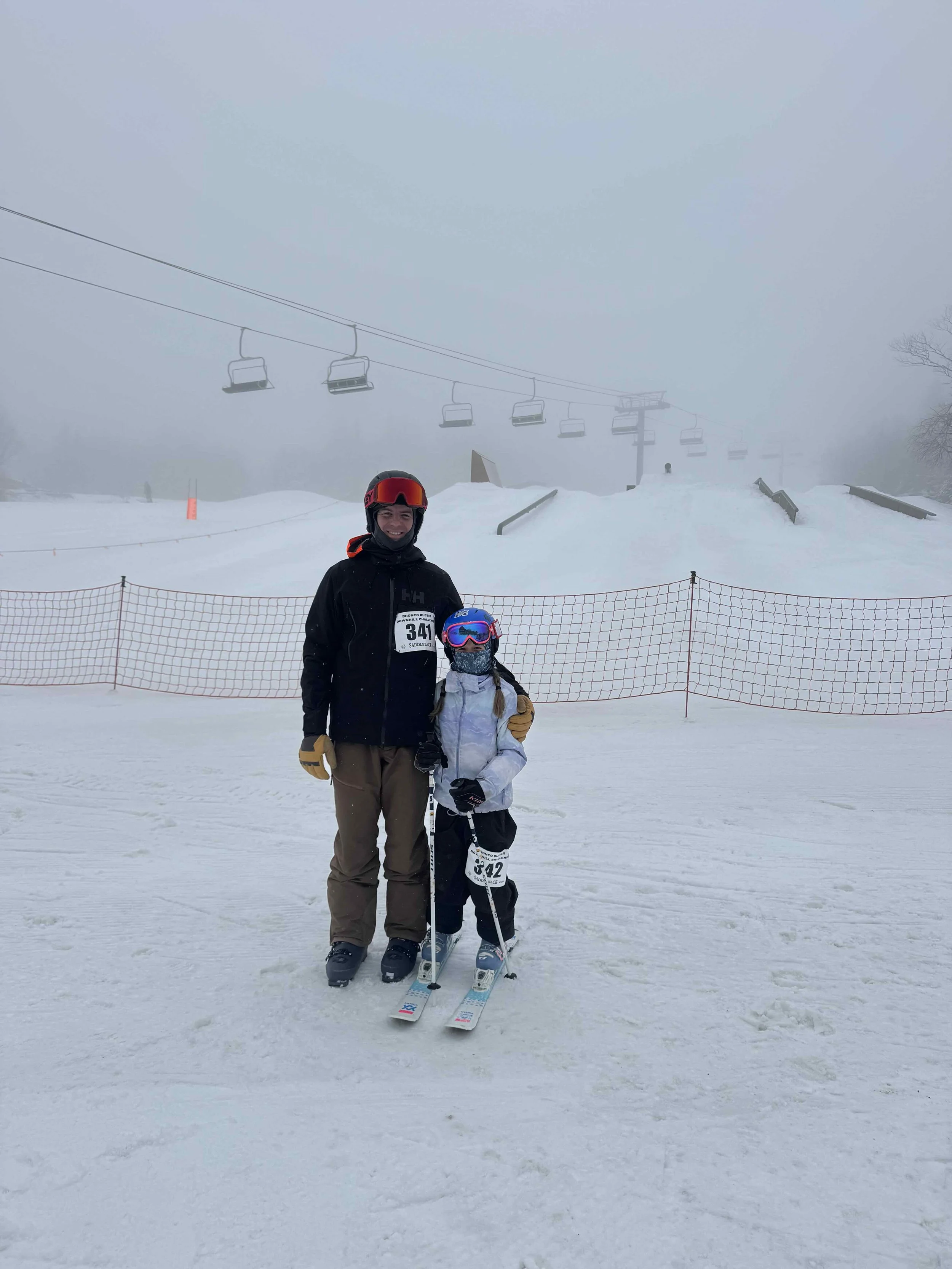 Two people standing on snow with ski equipment, wearing helmets and goggles, in front of a ski slope with a chairlift and foggy weather.