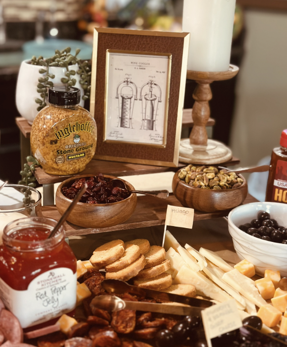 A cheese and charcuterie board with various cheeses, dried fruits, and nuts. There are bowls of dates, pistachios, and black olives. A jar of red pepper jelly and a bottle of honey are also present. In the background, a framed diagram of a device, a candle, and a potted succulent plant are visible.