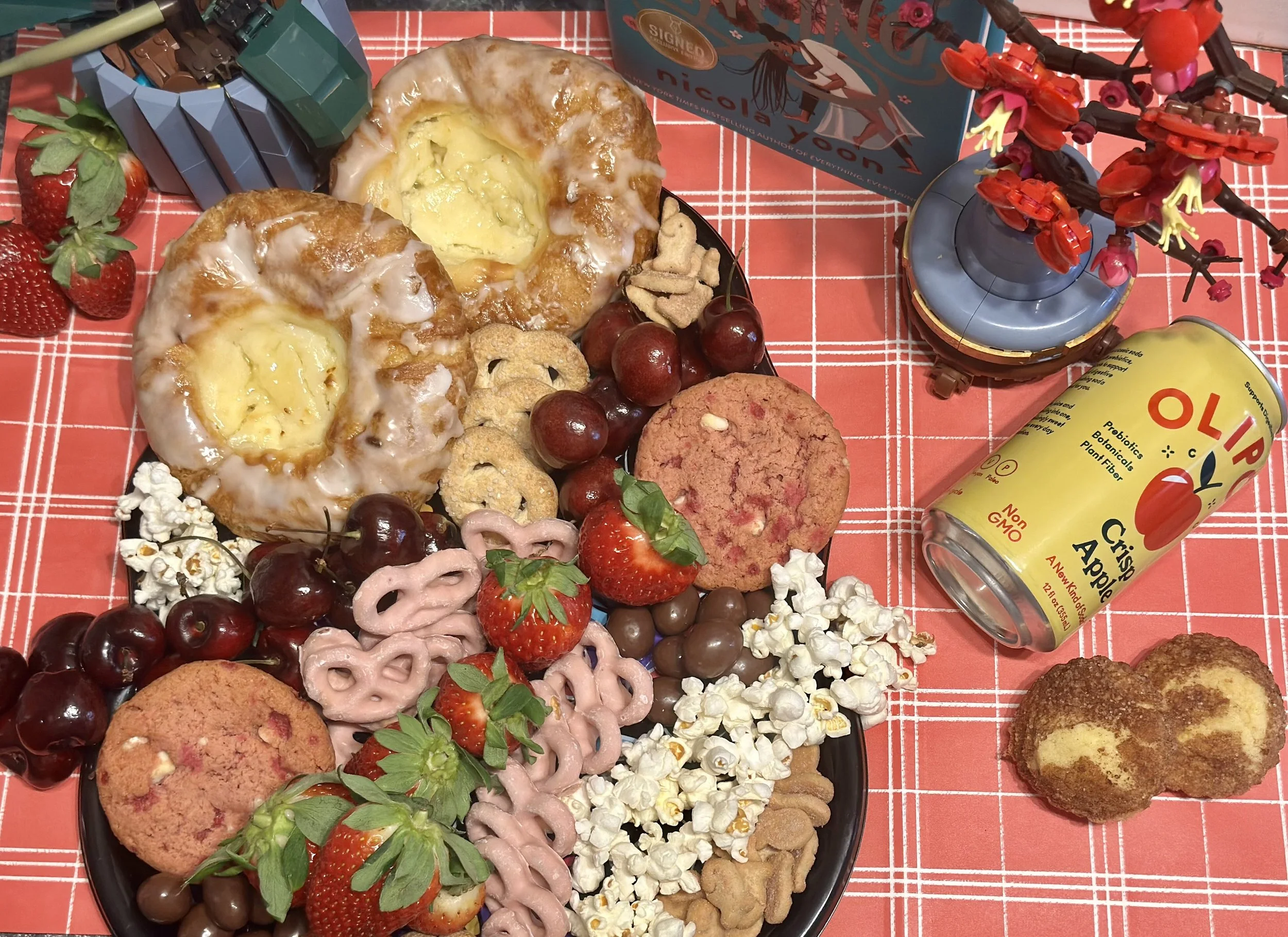 Charcuterie tray  with danishes, cherries, pink pretzels, pink cookie, strawberries, popcorn and more. With a yellow can drink that has a red apple in the center to the right Lego flowers on the left and right with a teal book in the back with a whit