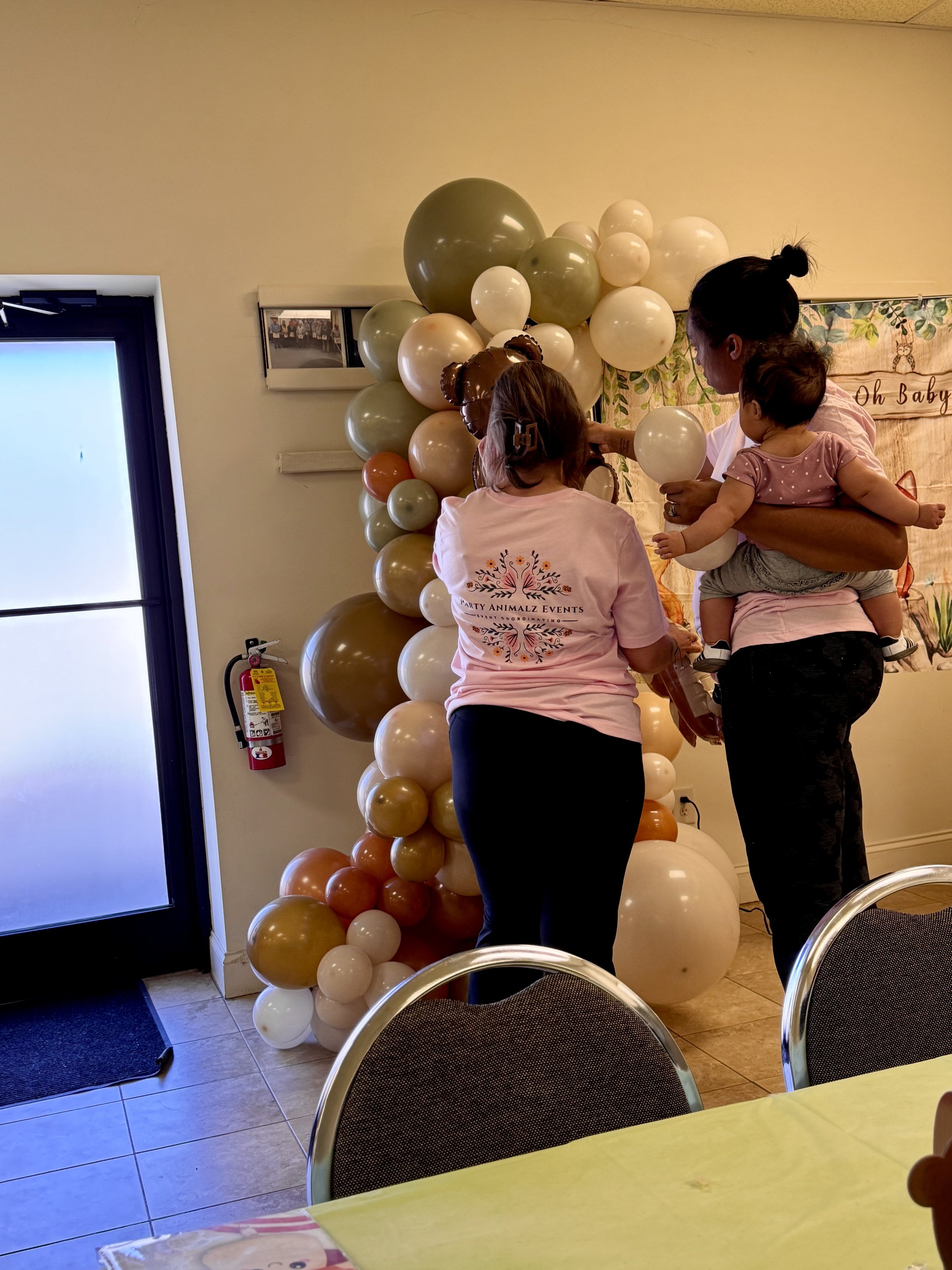 woodland themed balloon arch with animal balloons, two chairs and oh baby banner, two event worker with on holding a baby