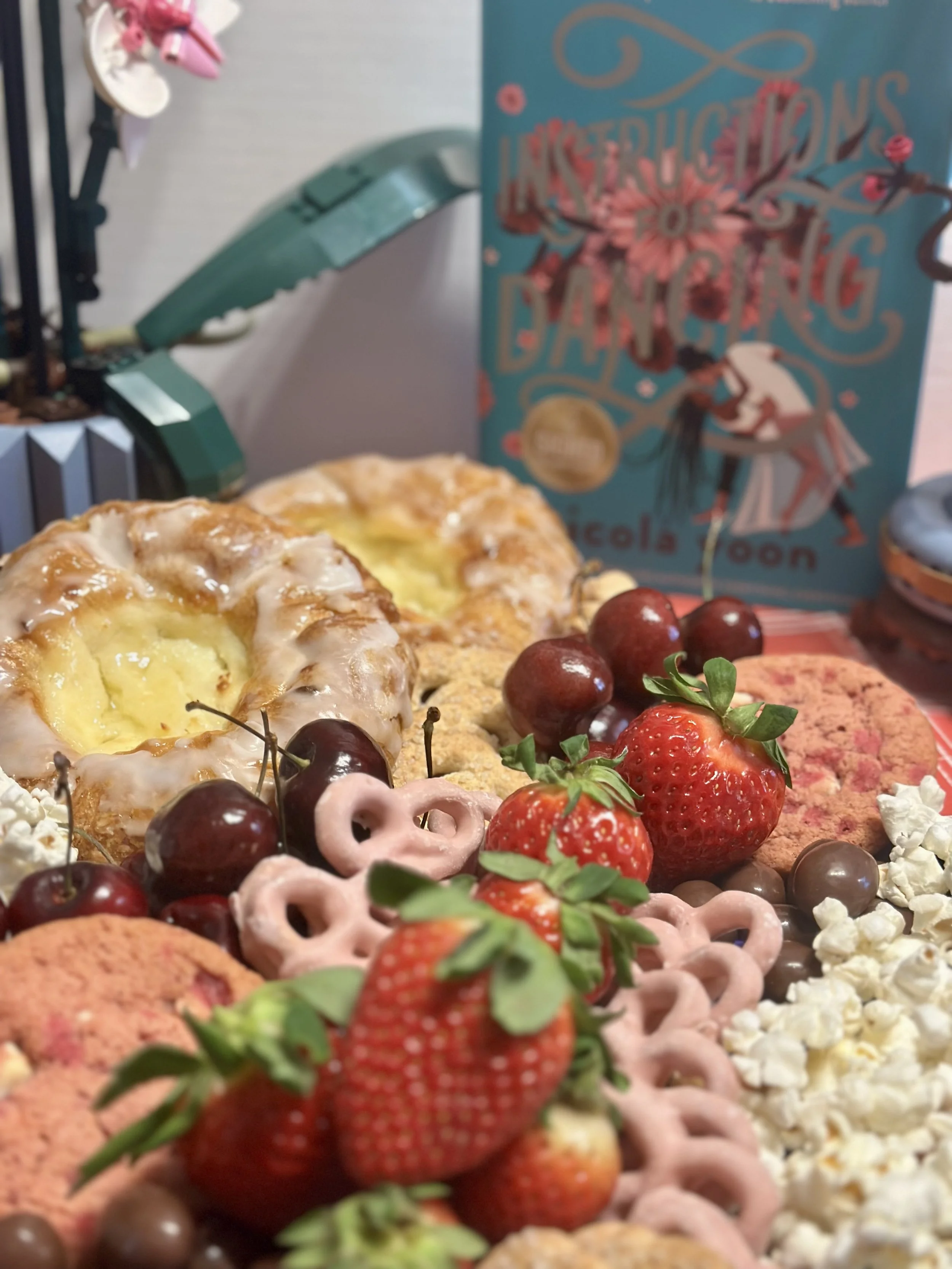 Charcuterie tray  with danishes, cherries, pink pretzels, pink cookie, strawberries, popcorn and more. Lego flowers on the left with a teal book in the back-drop