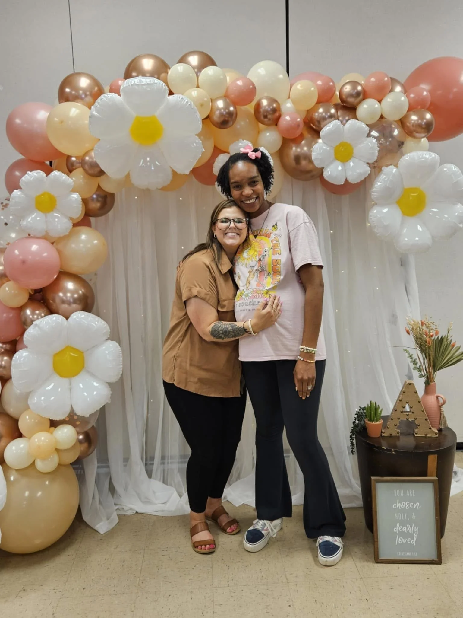 Two women standing in front of a balloon and fabric backdrop for a celebration. They are smiling and hugging. The background includes white, peach, and metallic balloons shaped like flowers, along with white sheer fabric. There is a small table with potted plants, a decorative letter, and a framed sign that reads 'You are chosen, holy and dearly loved.'