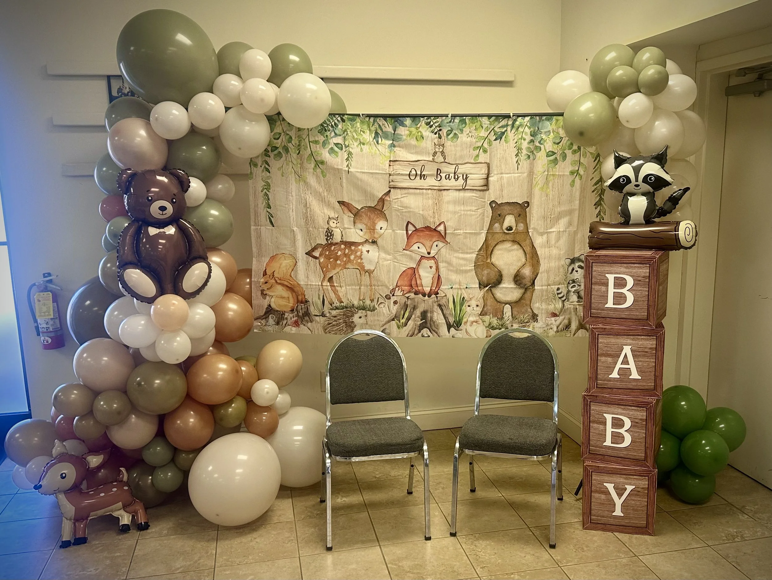 woodland themed balloon arch with animal balloons, wood-grained boxes spelling baby, two chairs and oh baby banner