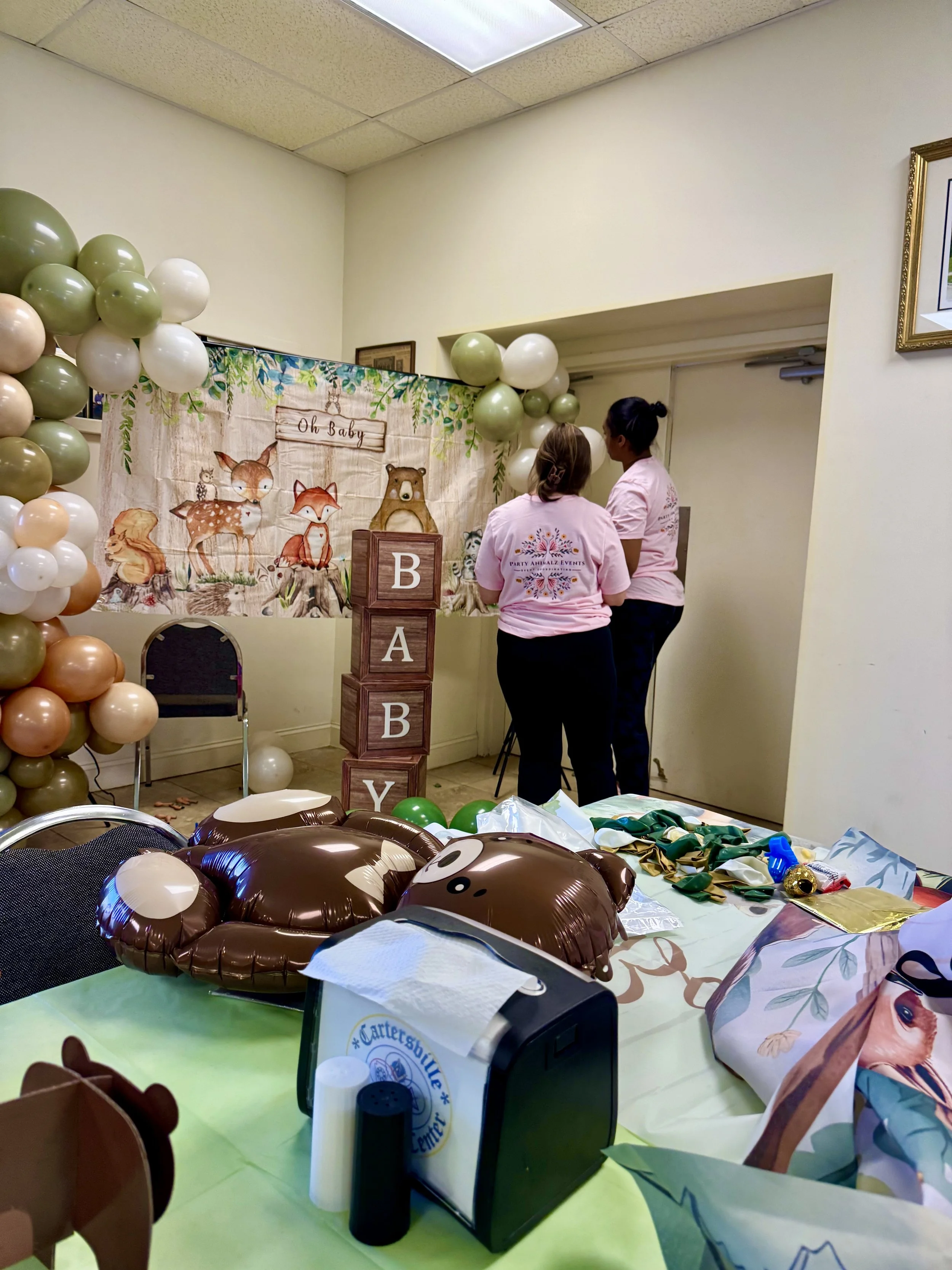 woodland themed balloon arch with animal balloons, top of a chair and party of the table with balloon supplies, oh baby banner, two event workers