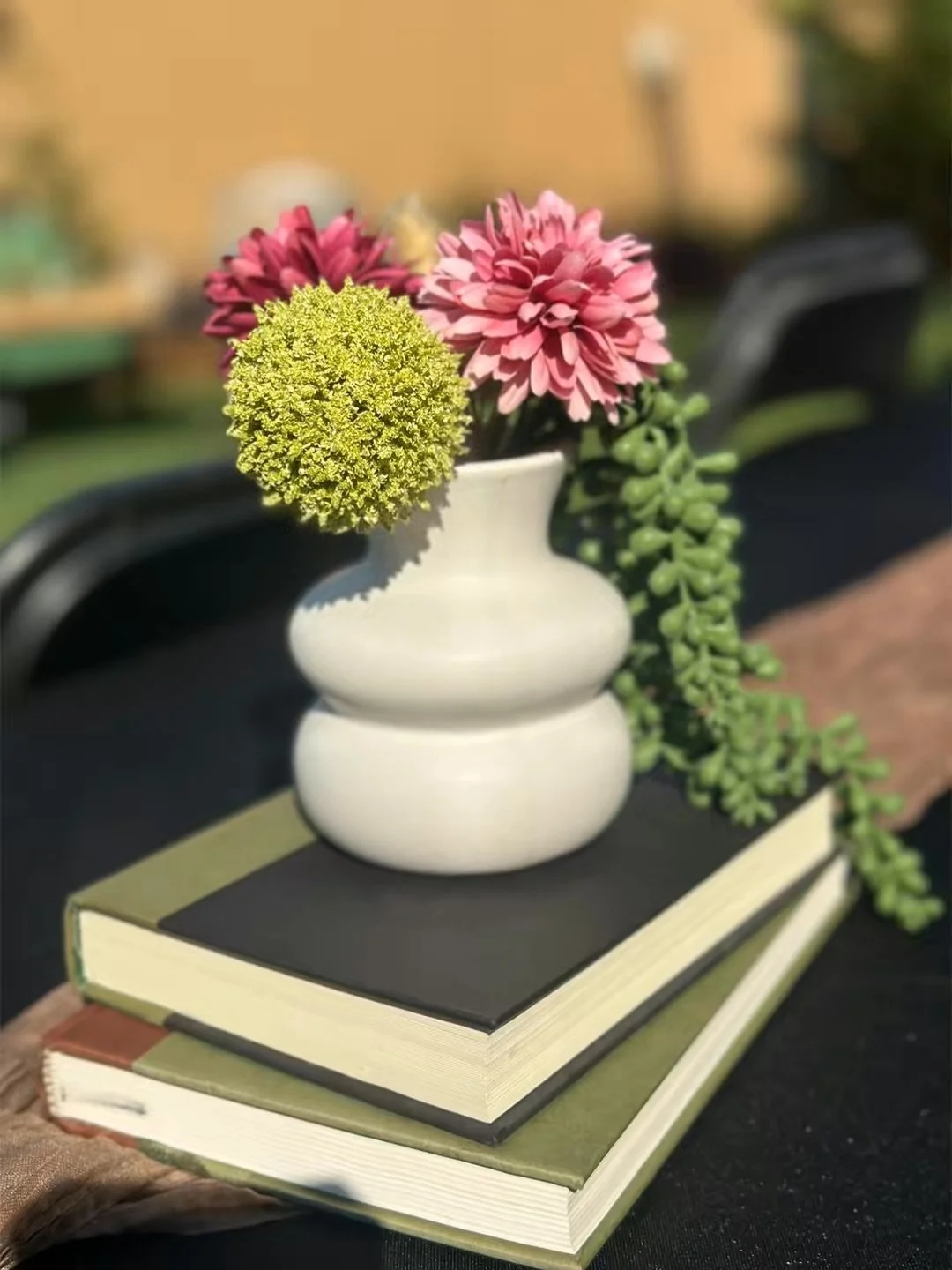 Pink and purple flowers in a white vase placed on two closed books, with one book being larger and green, and the other smaller and black, on a dark surface.
