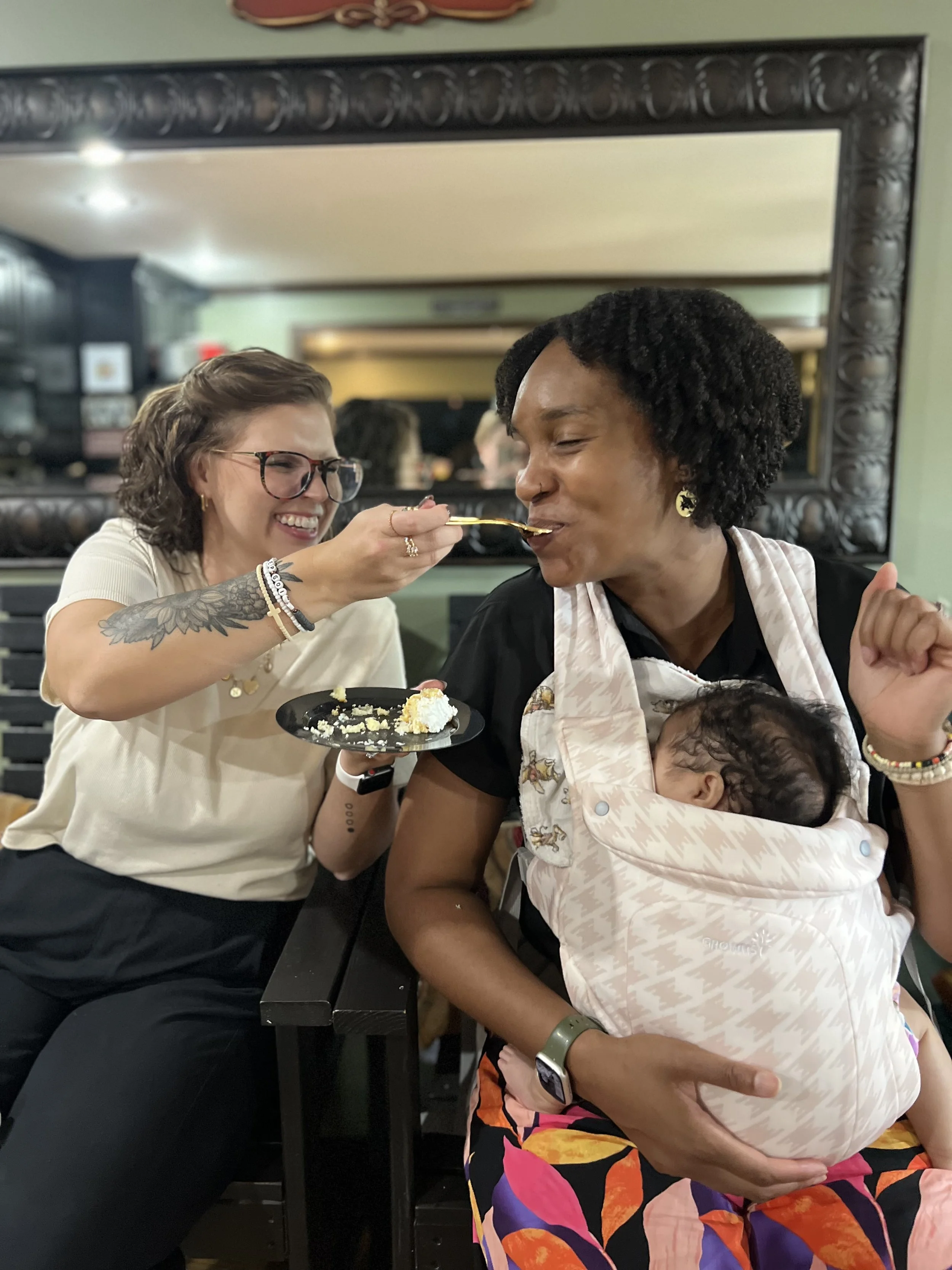 A woman with glasses feeding a piece of cake to a woman with curly hair wearing a baby carrier. The woman with glasses is smiling, and the woman with curly hair is closing her eyes as she eats. A baby is asleep in the carrier. They are in a restaurant with a large mirror reflecting the background.