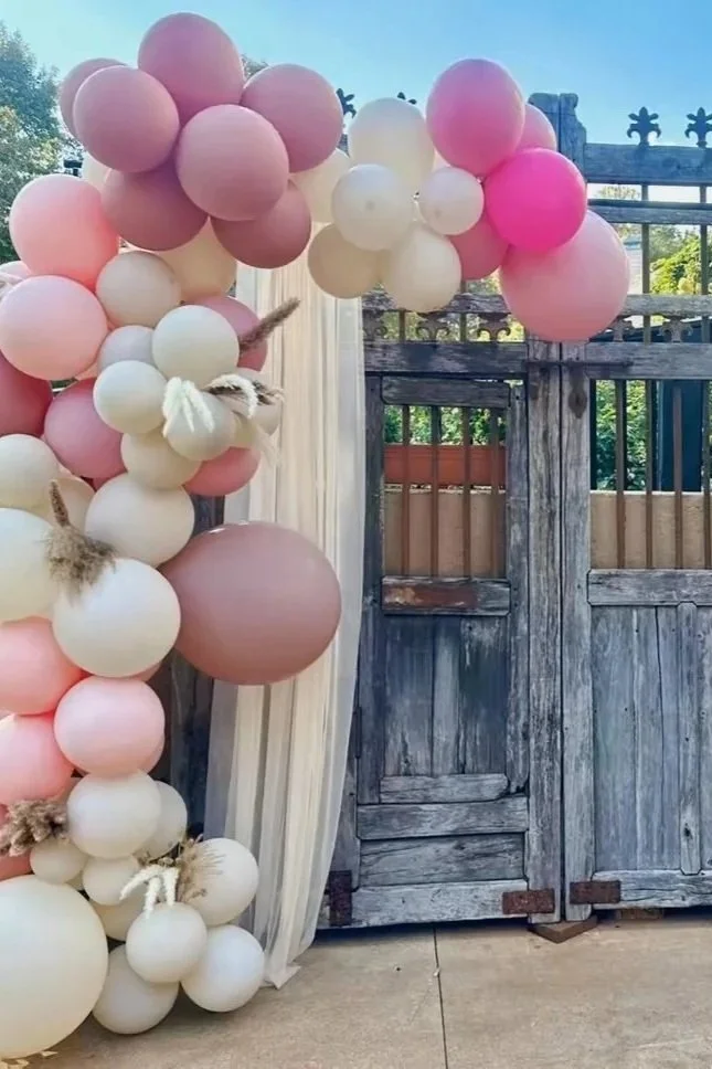 Colorful balloon arch with pink, cream, and peach balloons and decorative yarn knots, set against a rustic wooden gate and weathered fence outside.