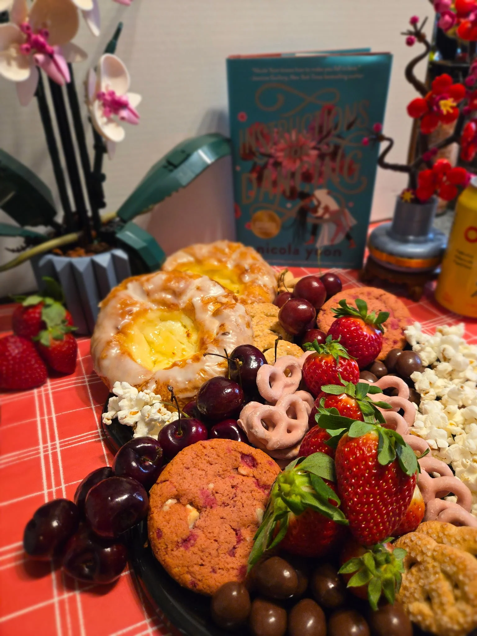 Charcuterie tray  with danishes, cherries, pink pretzels, pink cookie, strawberries, popcorn and more. Lego flowers on the left and right with a teal book in the back-drop