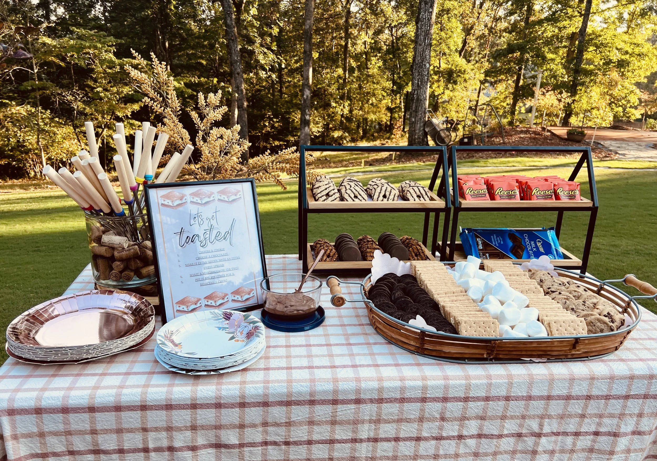 Outdoor dessert table with marshmallows, cookies, and chocolate bars, set on a checkered tablecloth in a backyard, with trees and sunlight in the background.