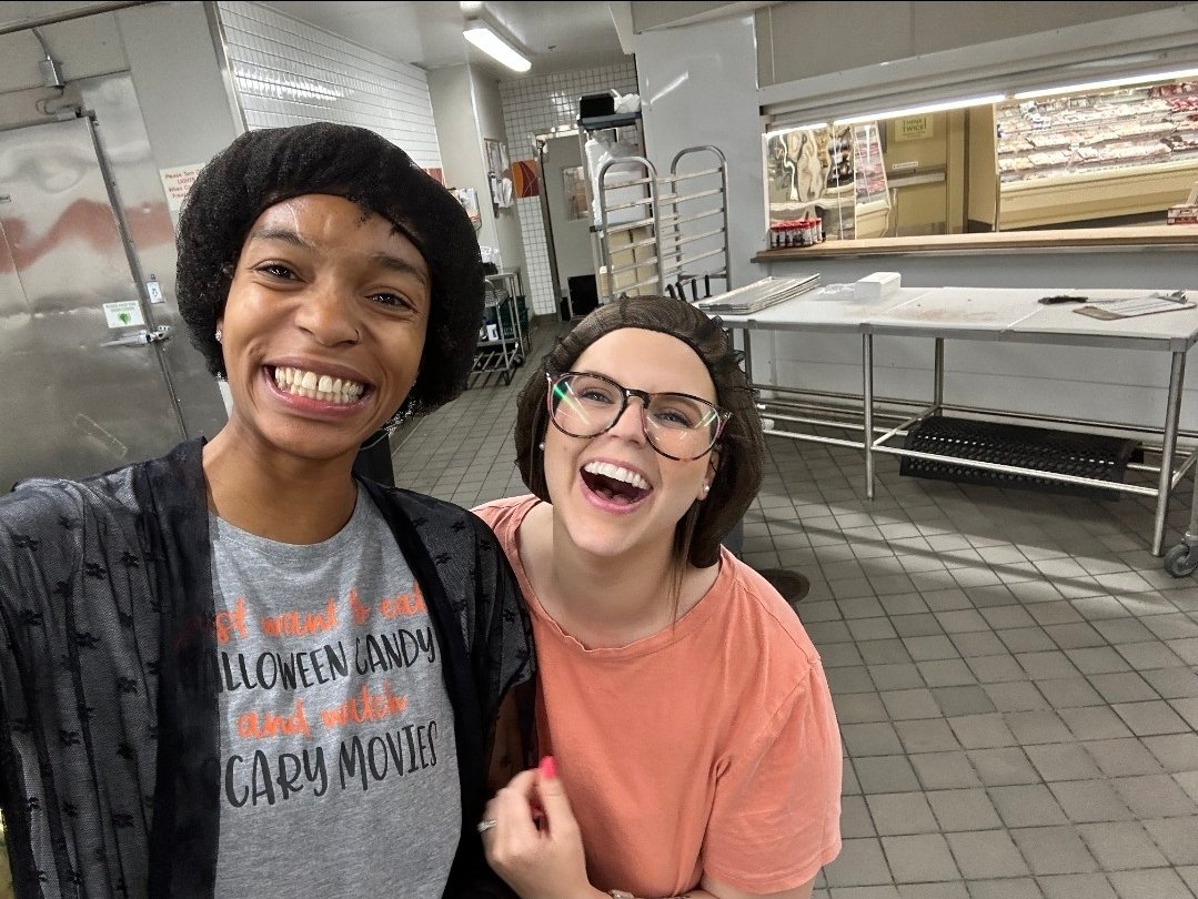 Two women smiling and taking a selfie in a commercial kitchen or bakery, with stainless steel countertops, trays, and kitchen equipment in the background.