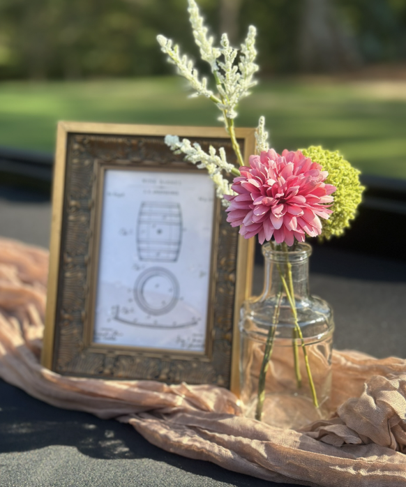 A decorative setup with a small bouquet of pink, green, and white flowers in a glass jar, a framed picture of a wine barrel, and a soft, beige fabric on a surface outdoors with greenery in the background.
