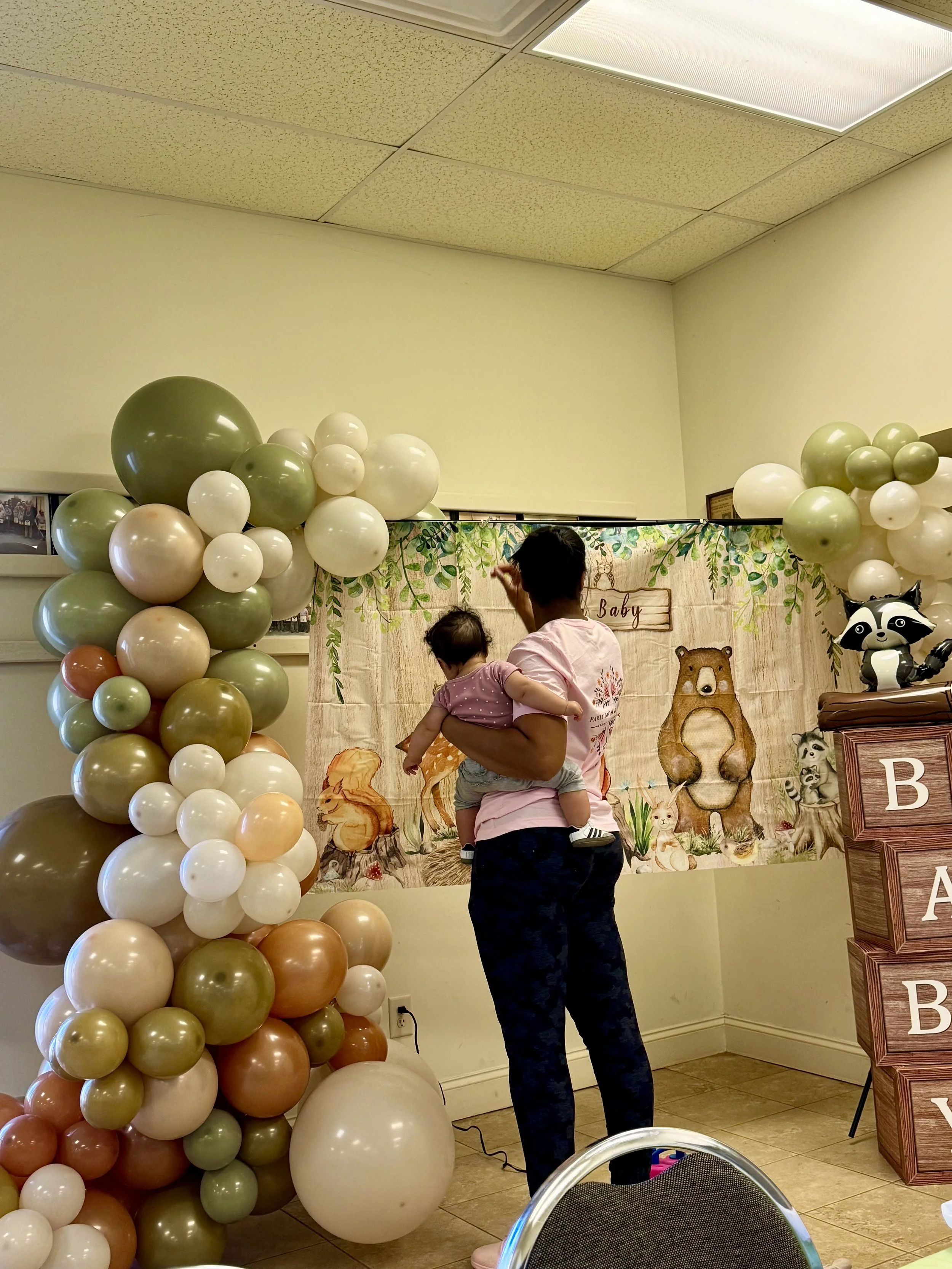 woodland themed balloon arch with animal balloons, wood-grained boxes spelling baby, top of a chair and oh baby banner, an event worker holding a baby
