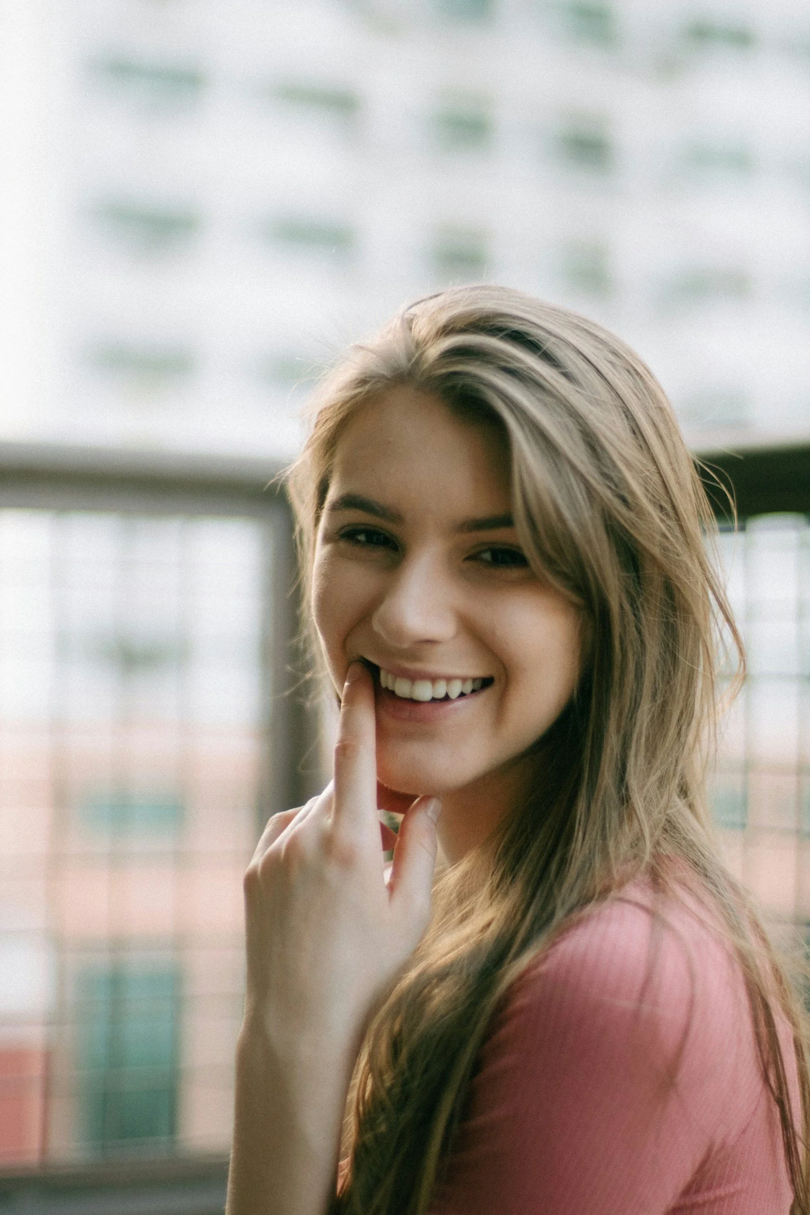 A smiling young woman with light brown hair and fair skin, wearing a pink top, standing outdoors near a blurred fence waiting for her personalized care appointment in Redmond Oregon