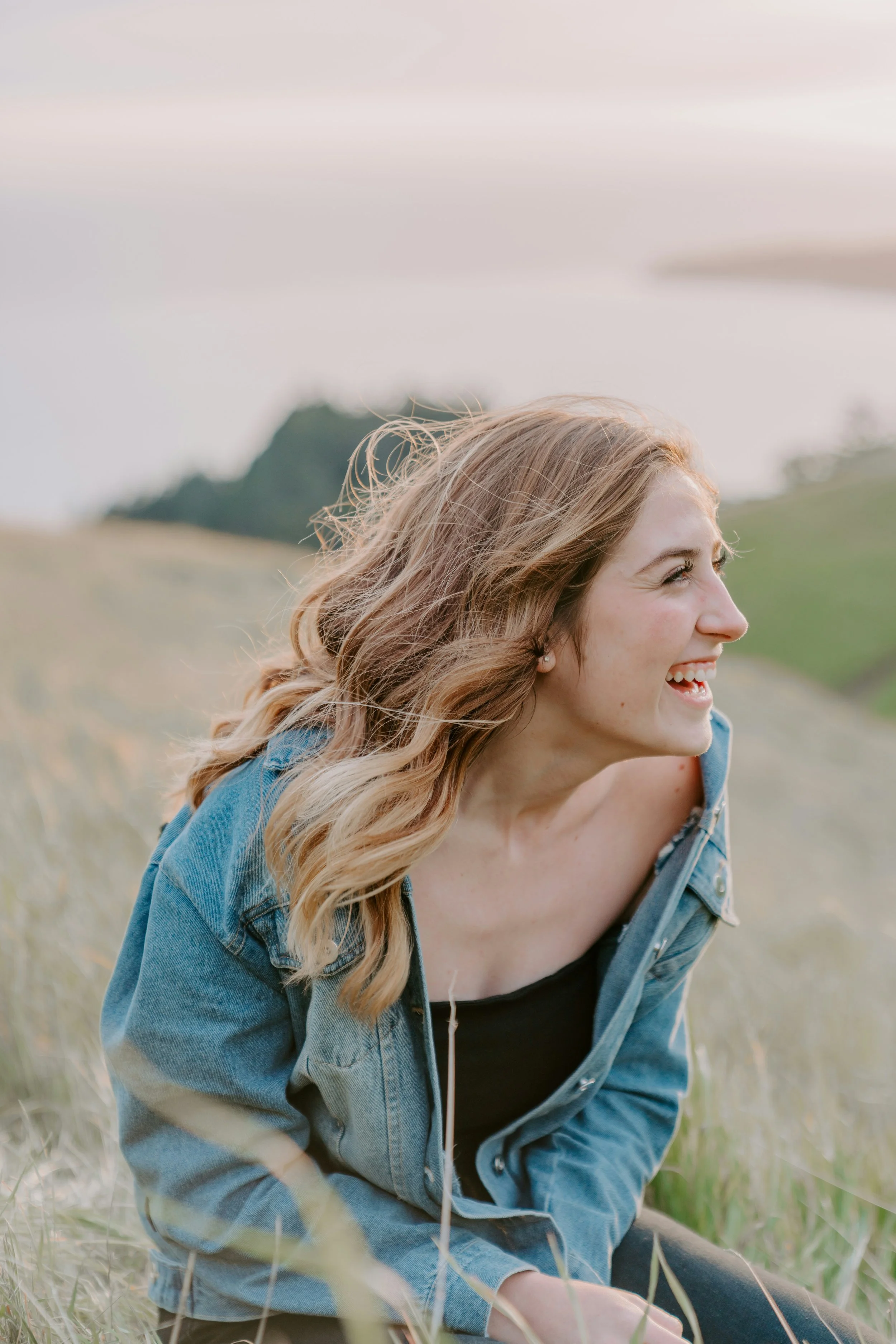 A young woman with long, wavy blonde hair, smiling and looking to her right, wearing a black top and blue denim jacket, sitting outdoors on grass. She represents that Alpine Wellness Direct Primary Care offer services to teenagers (14-18)